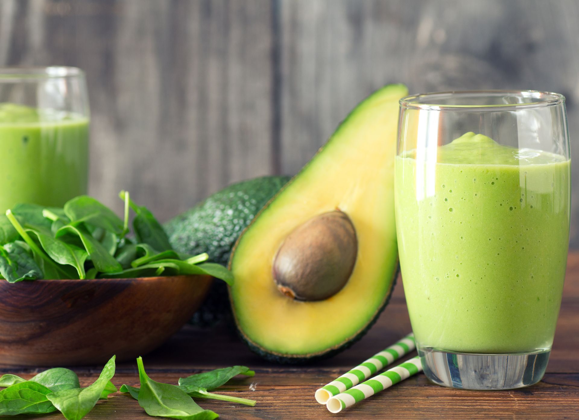 Green smoothie in glasses, alongside spinach, avocado, and straws, on a wooden surface.