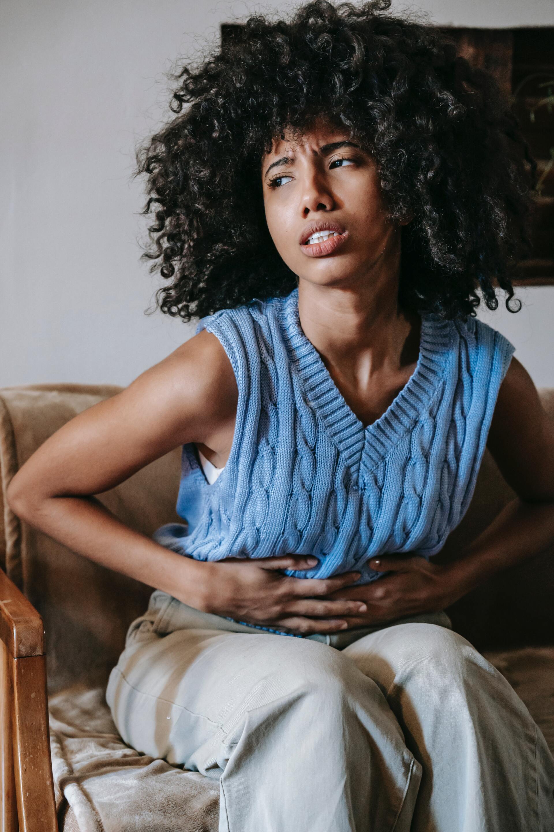 A woman with curly hair clutches her stomach while sitting on a sofa, showing an expression of pain. She wears a blue knit vest and light pants.