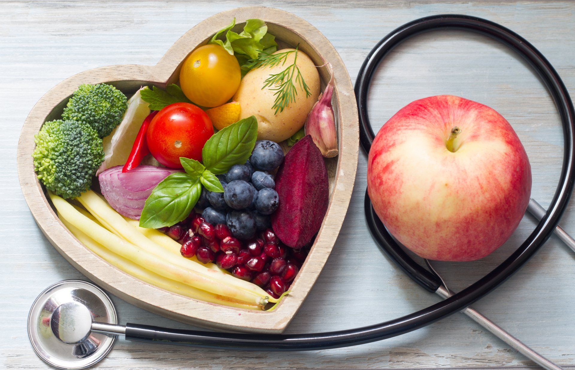 Heart-shaped bowl filled with colorful vegetables and fruits, next to an apple and a stethoscope, symbolizing heart-healthy diet.
