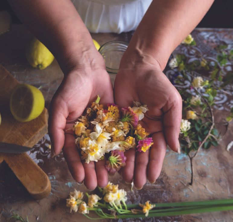 Woman's hands cupped, holding colorful flower petals. Lemons and a cutting board are visible on the rustic table.