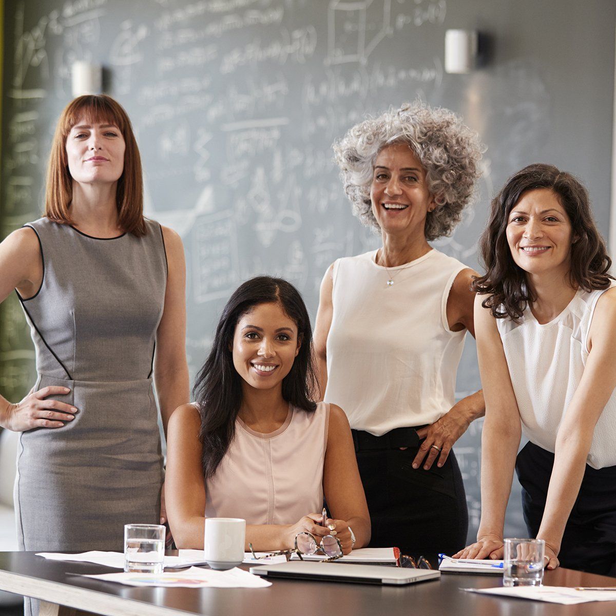 Four diverse women smiling around a table in a modern office. They appear to be in a business meeting, with a whiteboard visible in the background.