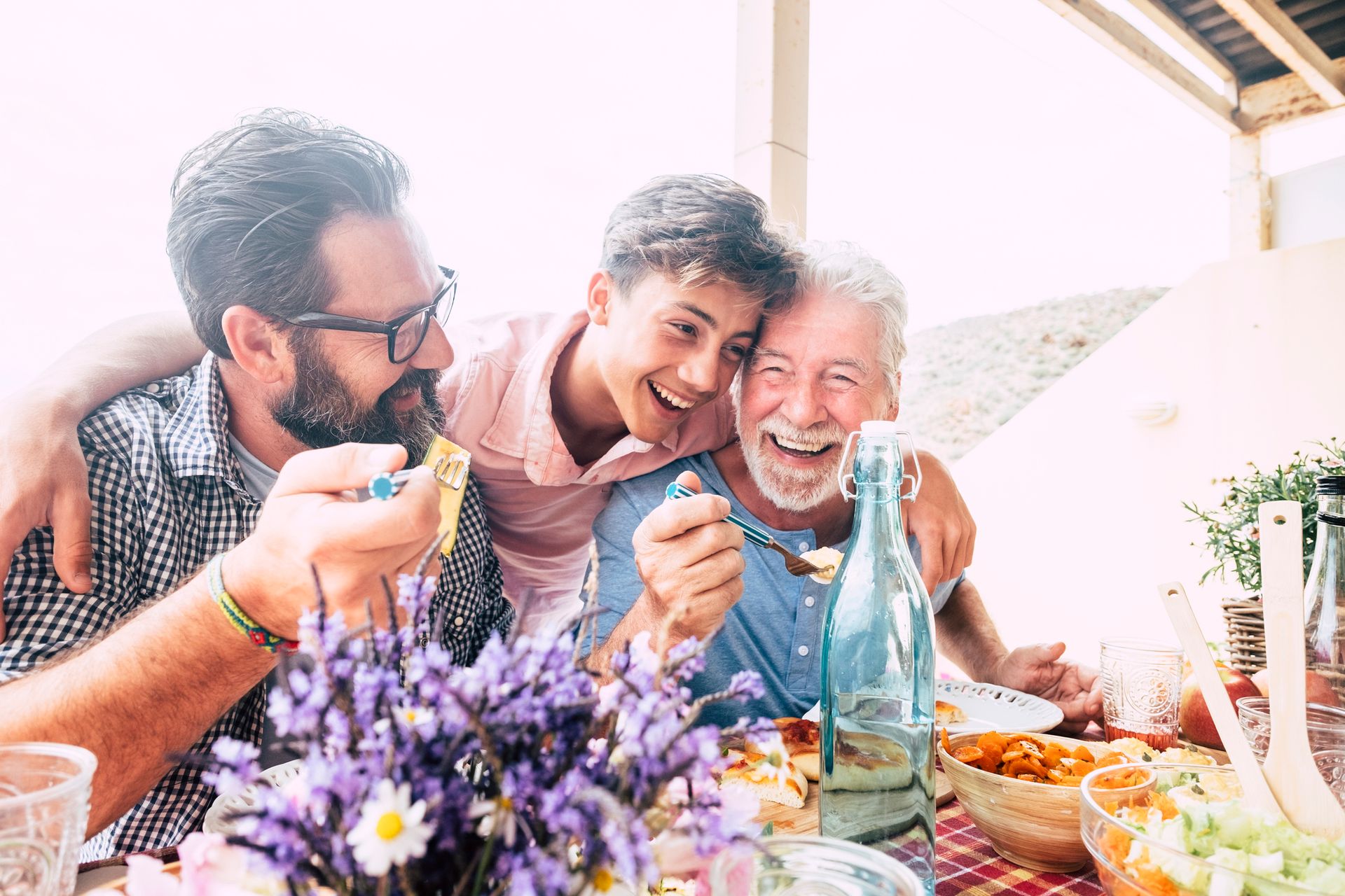 Three generations smiling and eating together at an outdoor table: a father with a beard, a teenager with his arms around the grandfather.