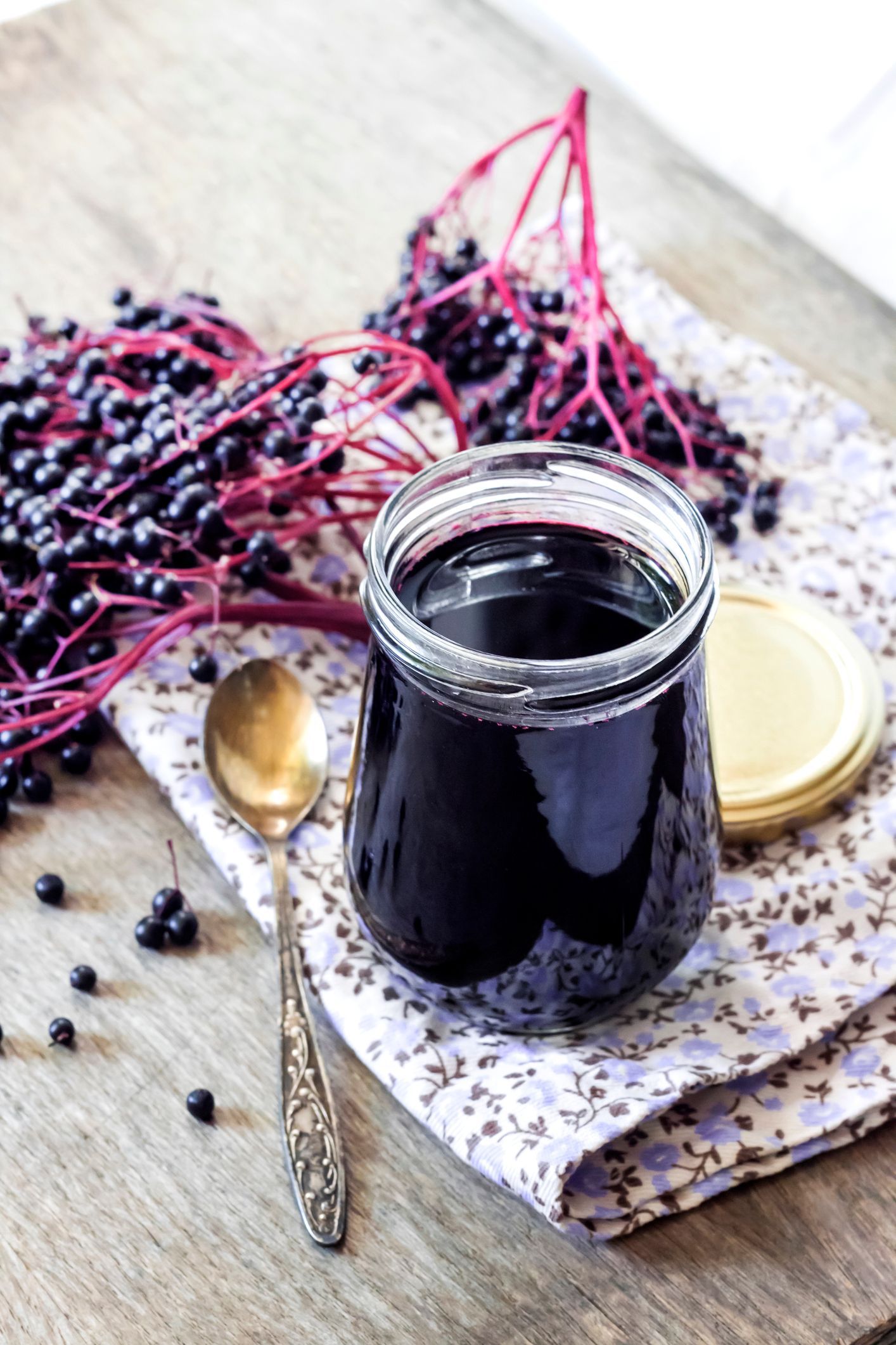 Jar of dark purple elderberry syrup, next to a spoon and elderberries on a floral napkin and wooden surface.