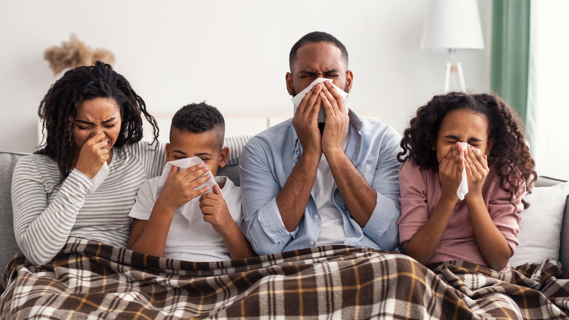 Family of four, sitting on a couch, each blowing their nose with a tissue; all look sick.