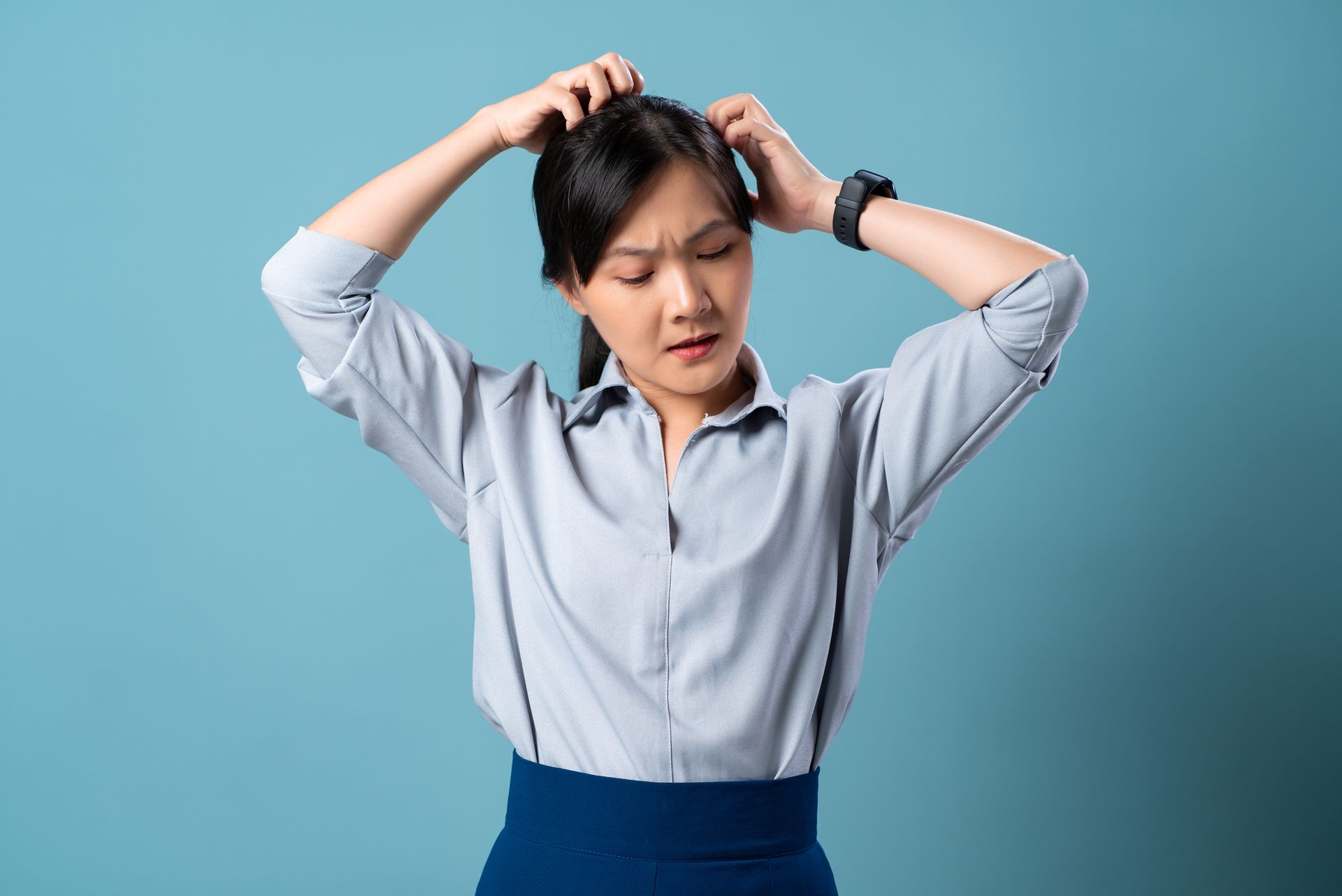 Woman in light blue button-down shirt scratching her head against a blue backdrop, looking confused.