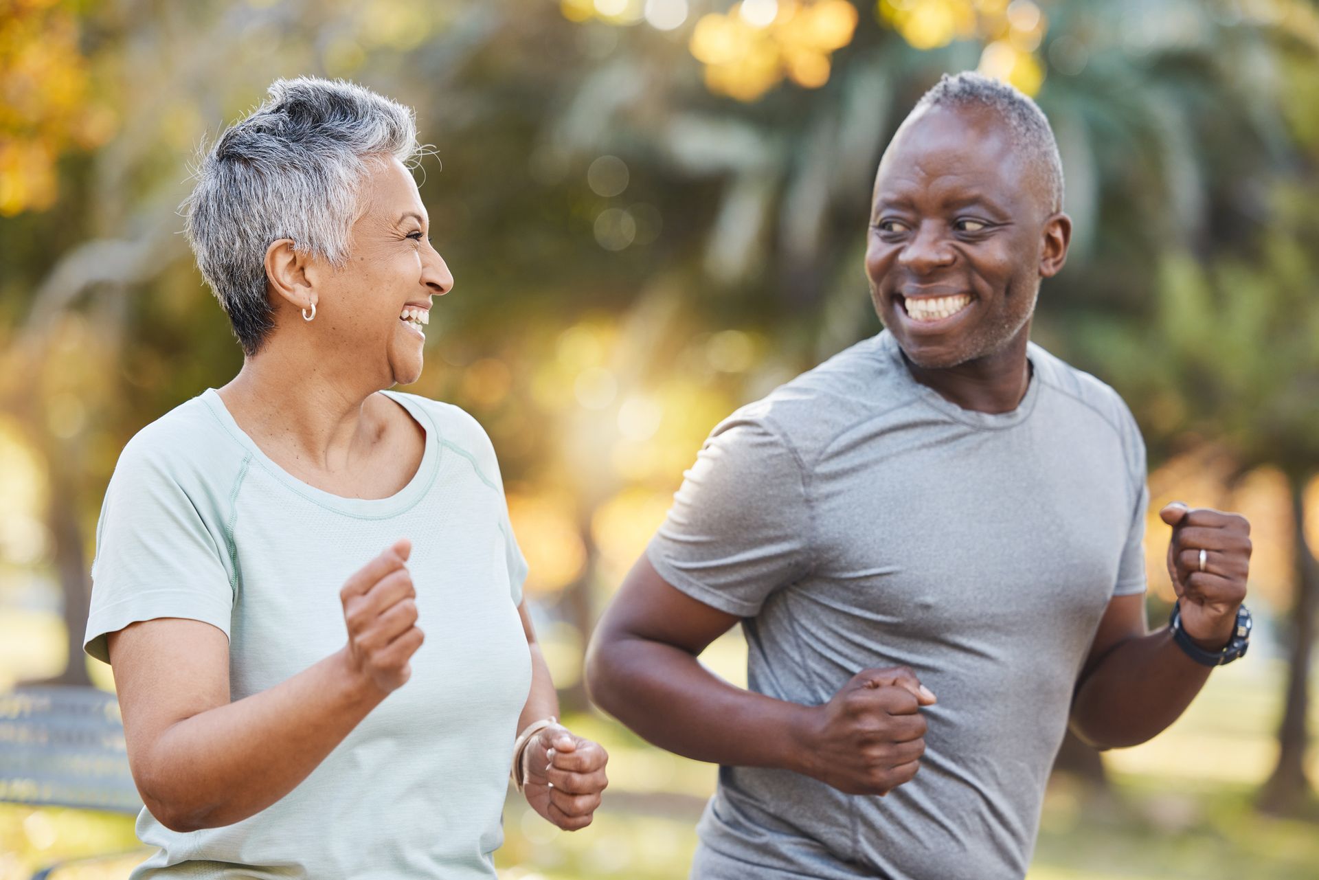 Smiling older couple jogging outdoors in a park; woman with gray hair and light blue shirt, man in gray shirt.