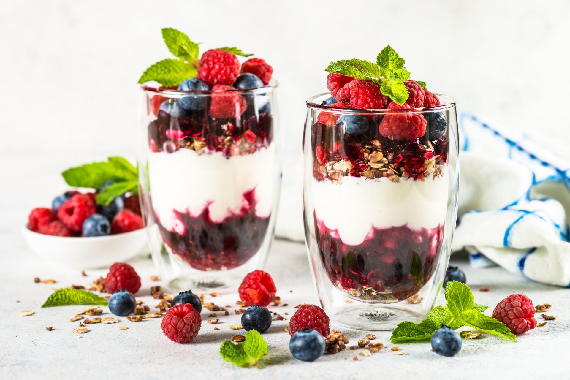 Two layered berry parfaits in clear glasses, topped with fresh berries and mint, with a bowl of berries nearby.