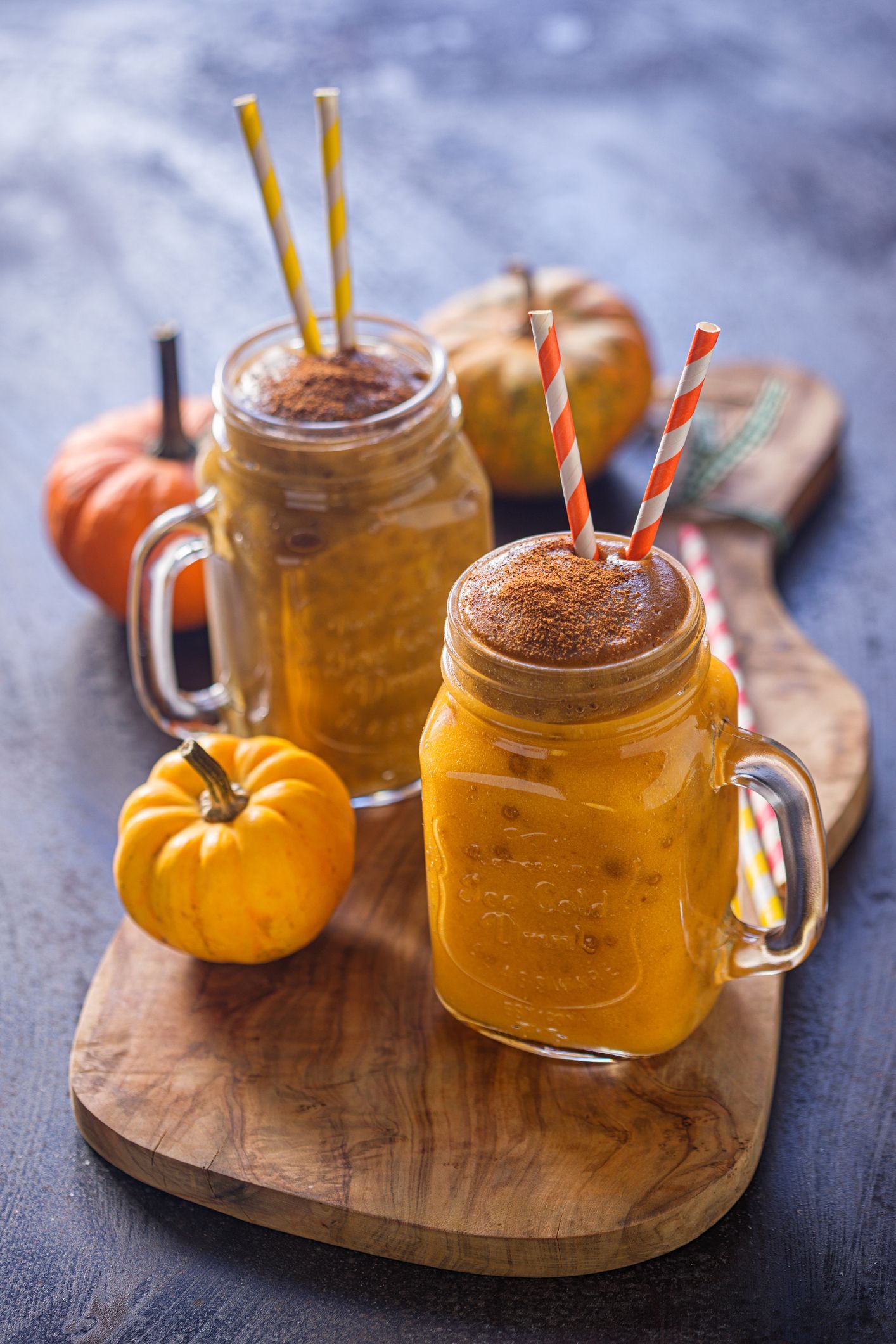 Two pumpkin spice smoothies in mason jars with straws on a wooden board, surrounded by mini pumpkins.