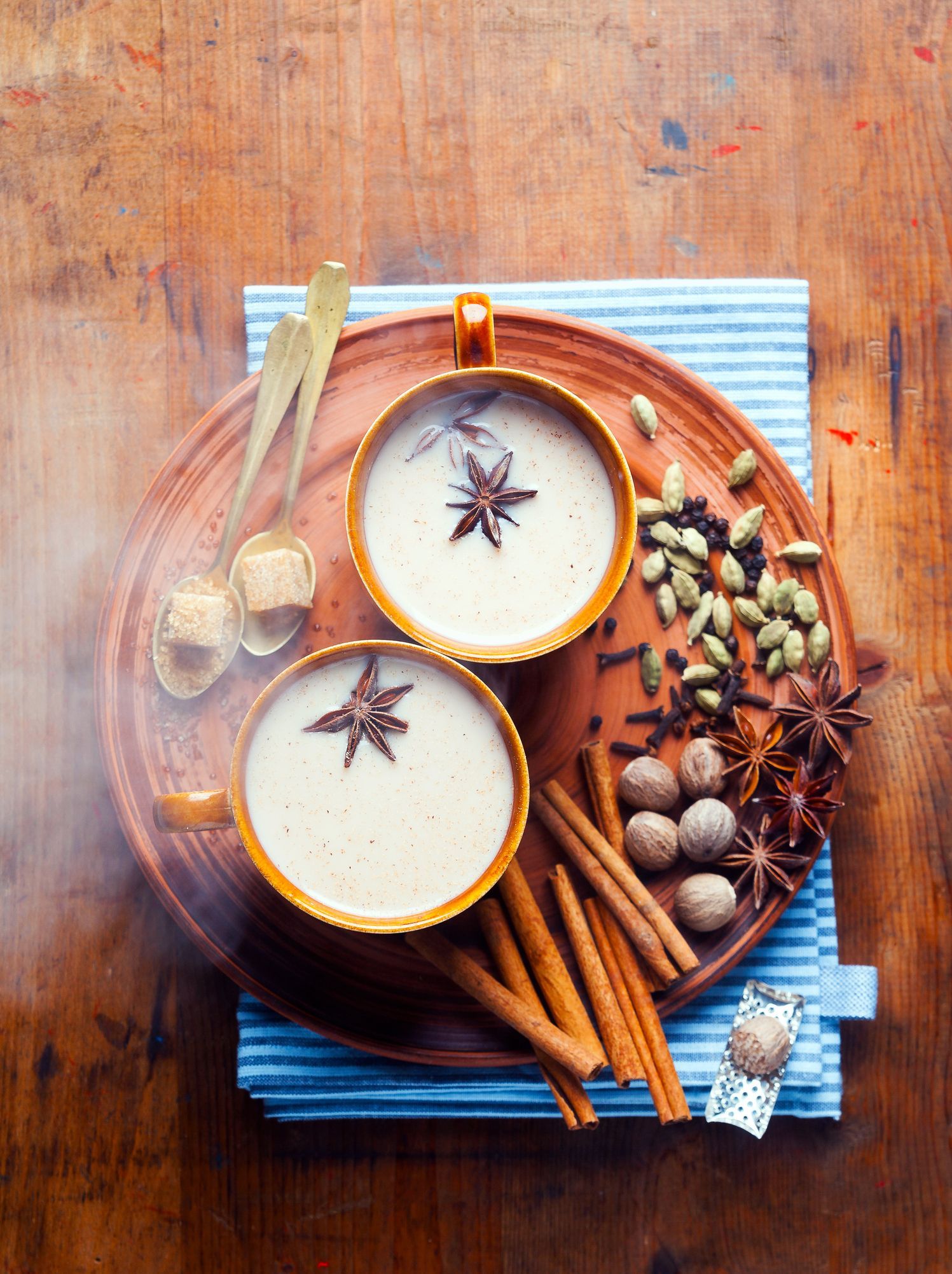 Two mugs of chai tea on a wooden tray with spices and sugar cubes.