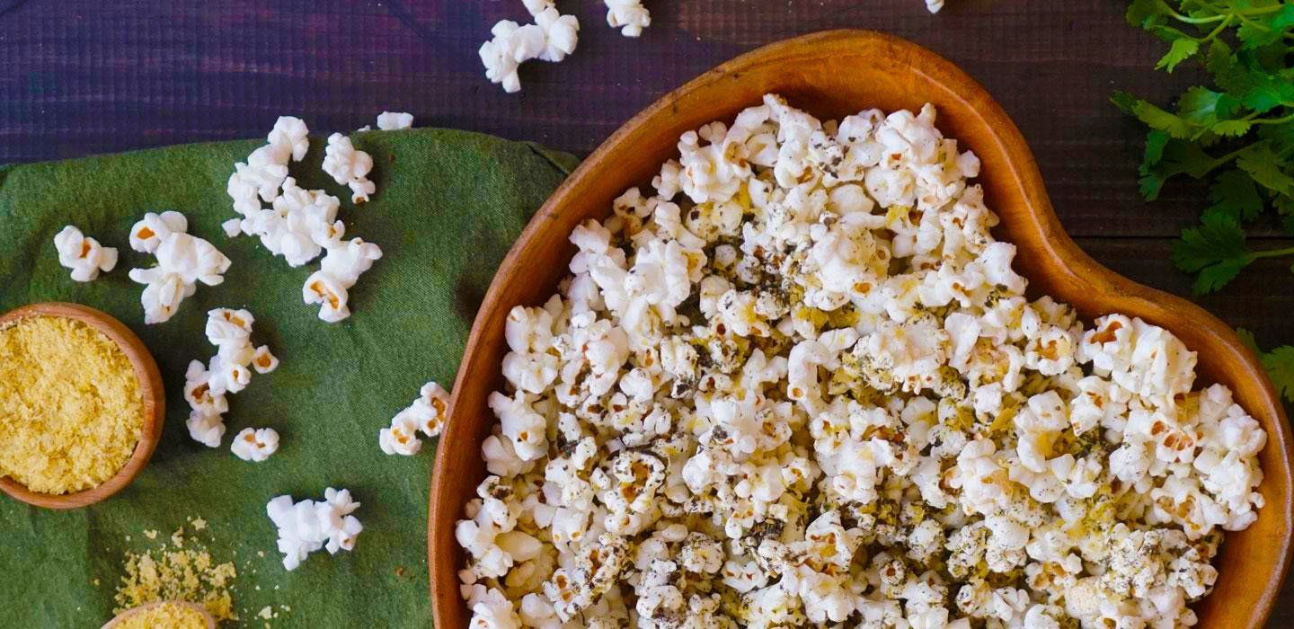 A wooden heart-shaped bowl filled with popcorn, next to a small bowl of yellow powder, and on a green cloth.