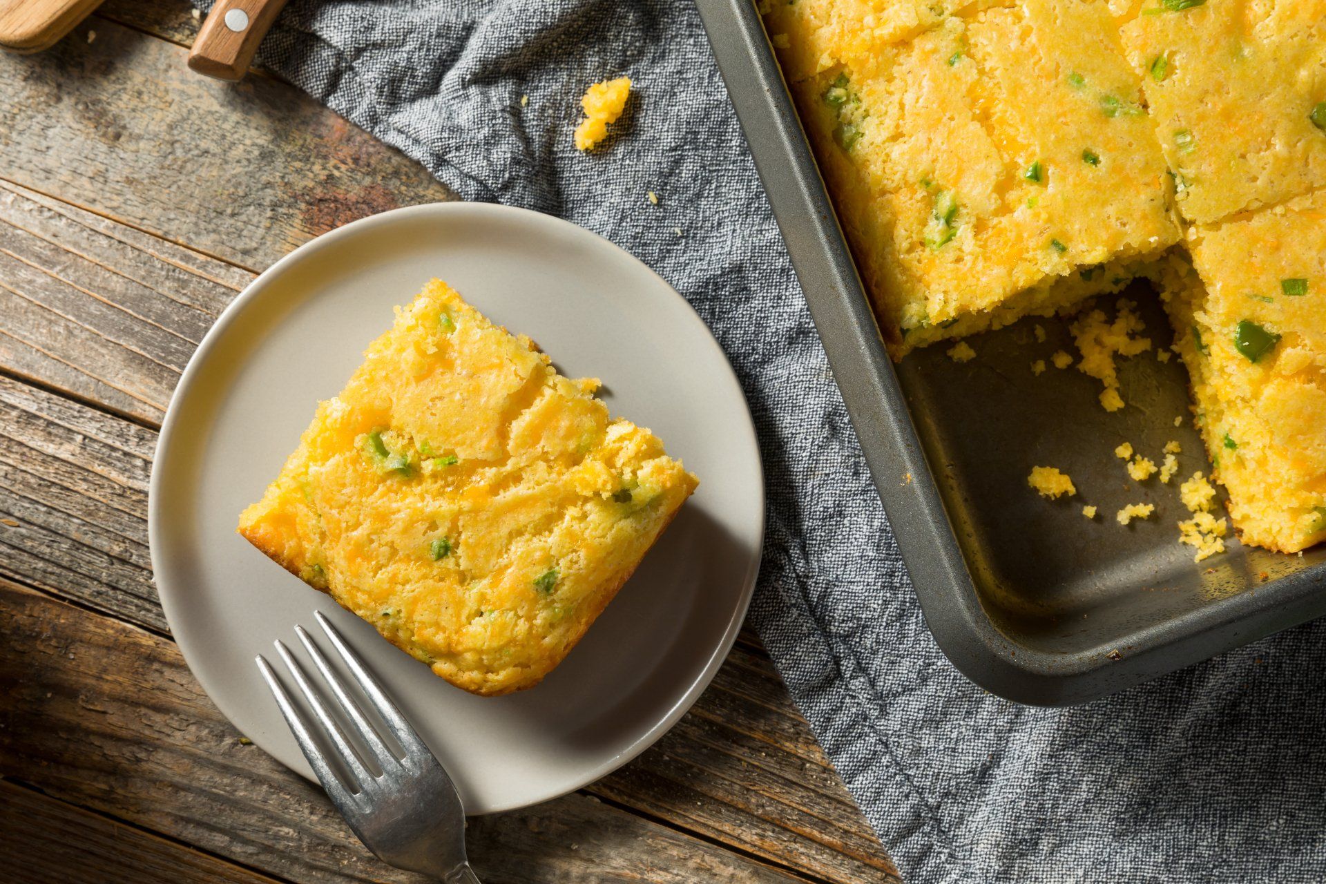 Cornbread slice on plate, next to pan of cornbread, on rustic wooden surface.