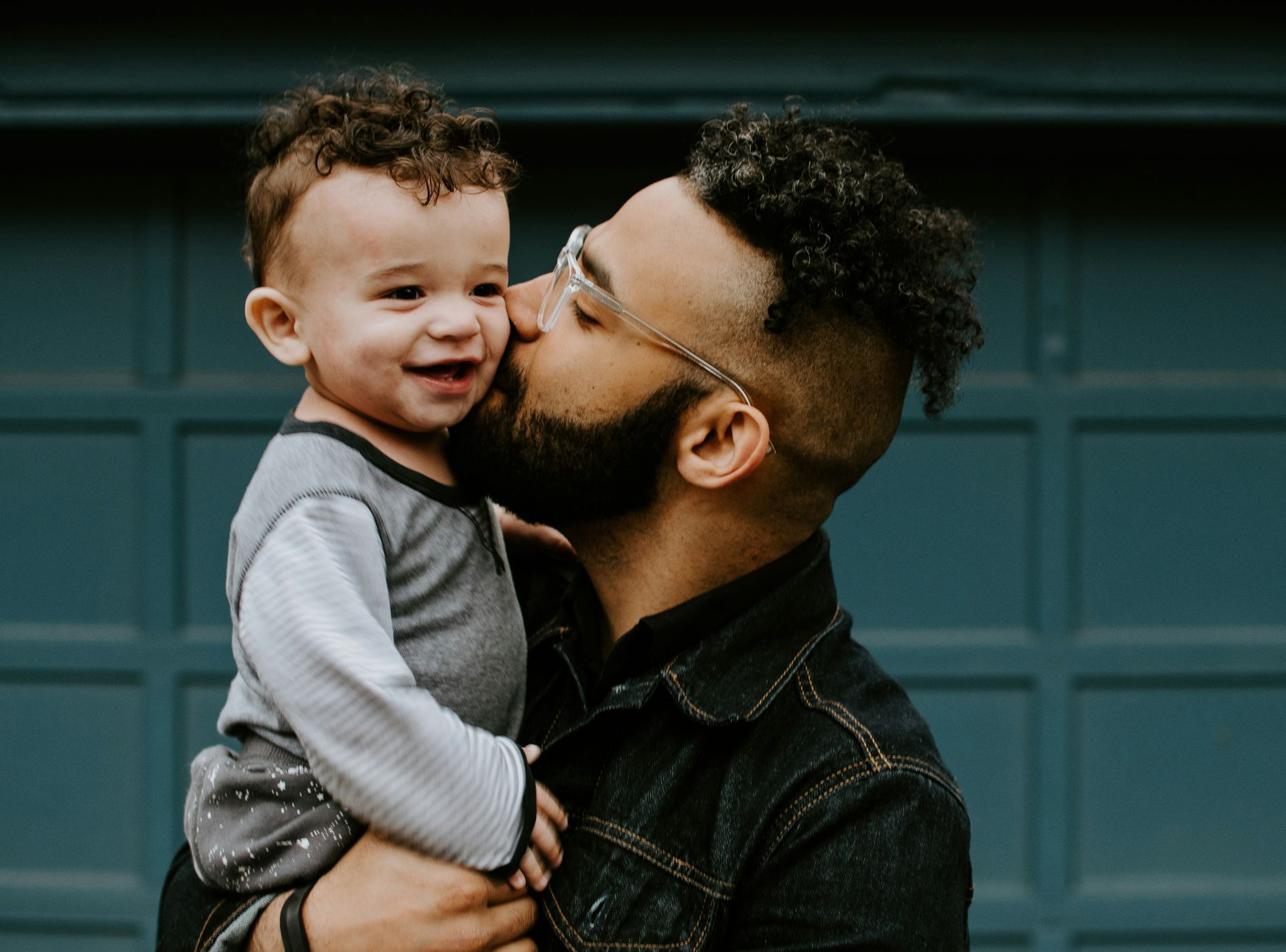 A man kisses a smiling baby on the cheek in front of a blue garage door. The man wears glasses and a denim jacket.