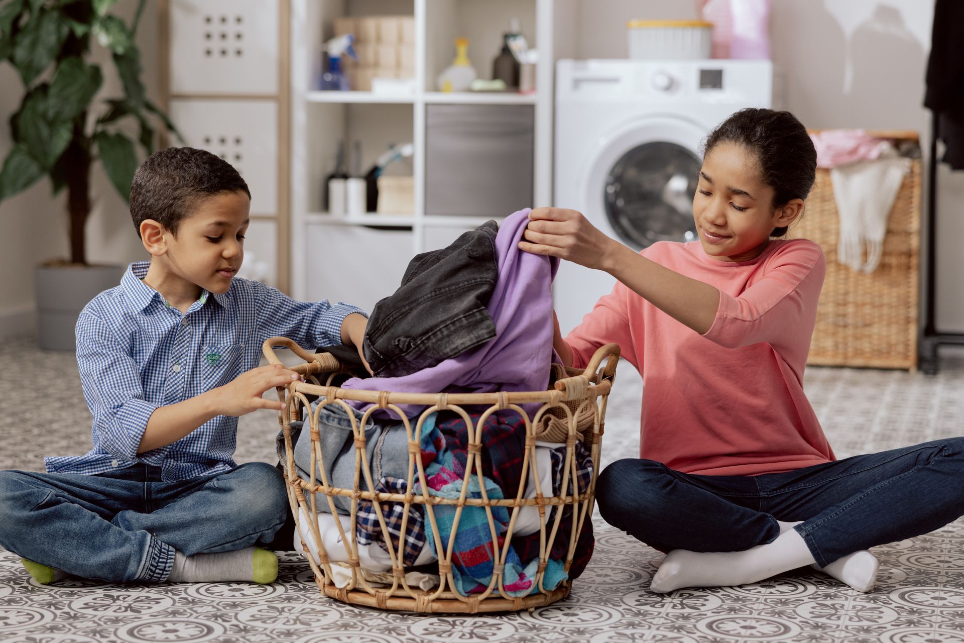 Two children sorting laundry from a wicker basket on a rug. The girl holds a purple shirt while the boy looks on. A washing machine is in the background.
