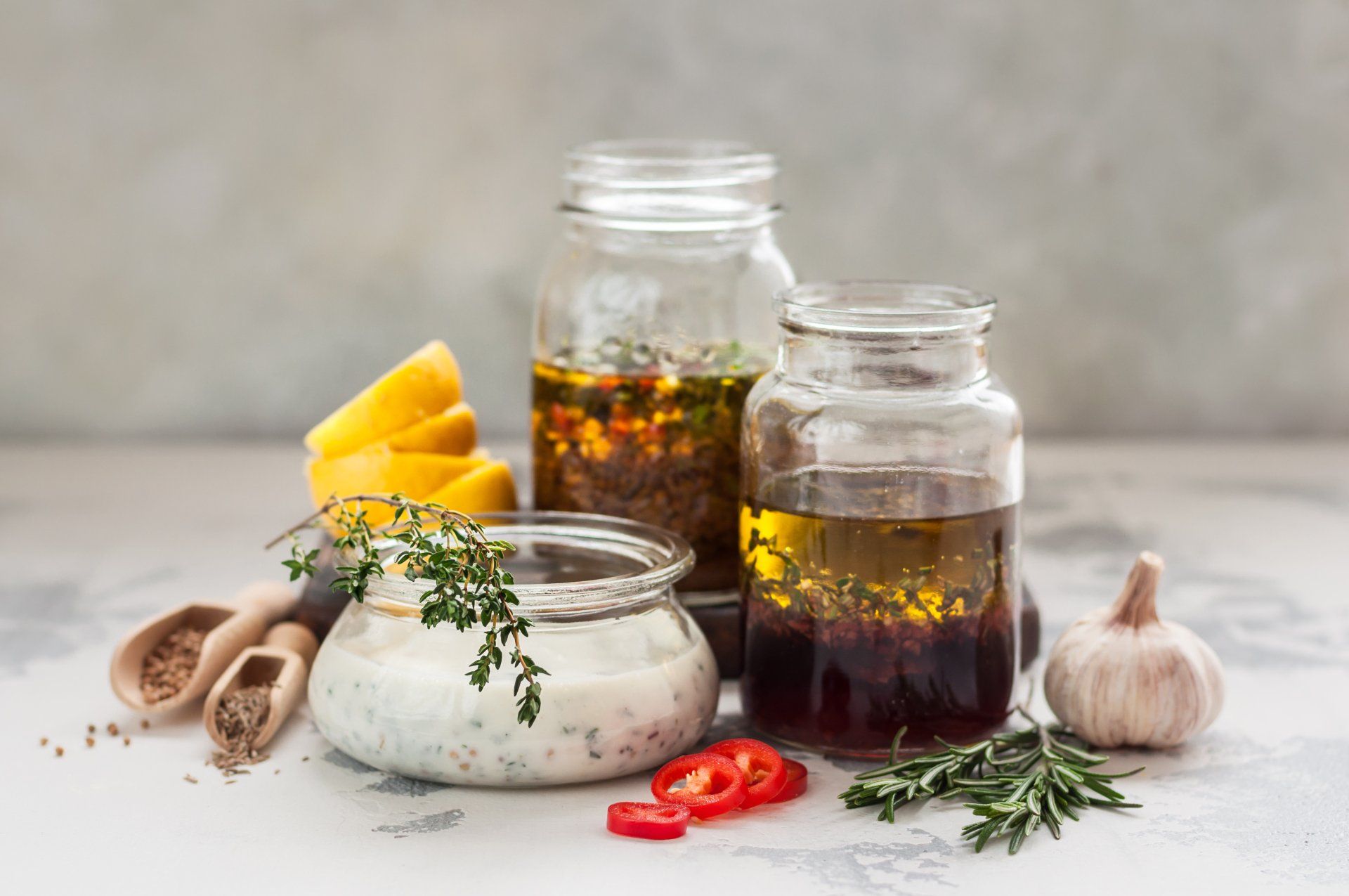 Jars of homemade salad dressings with ingredients like lemon, garlic, herbs, and chili peppers arranged on a light surface.