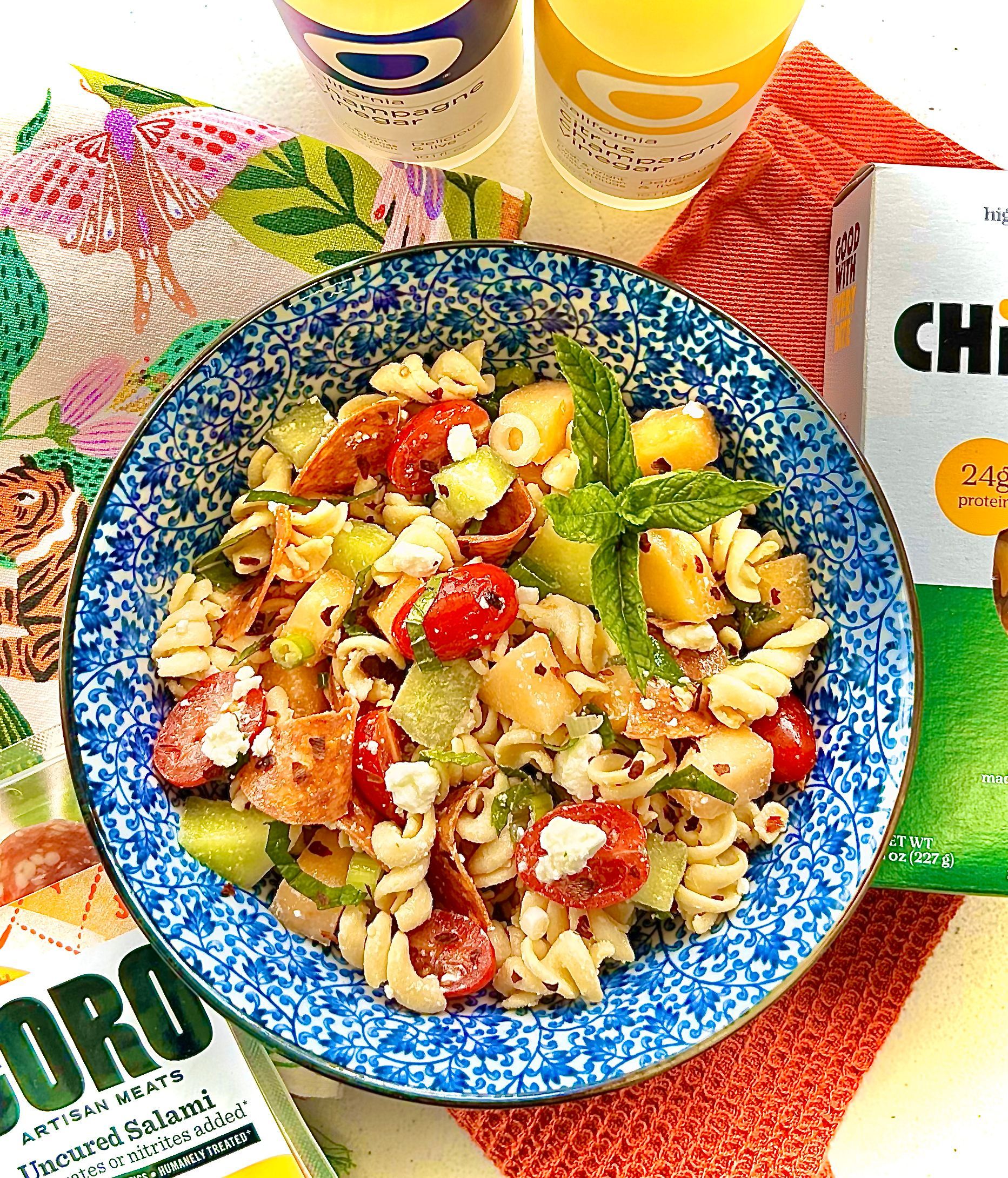 Pasta salad with tomatoes, cucumbers, and feta cheese in a blue bowl, next to drinks and chips.