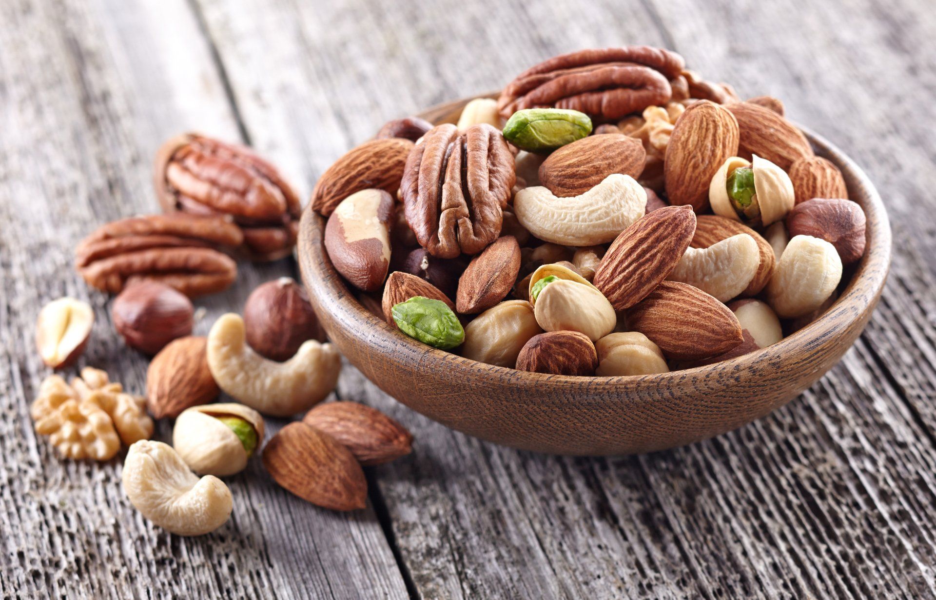 Bowl of mixed nuts on a wooden surface: pecans, almonds, cashews, pistachios, and walnuts.