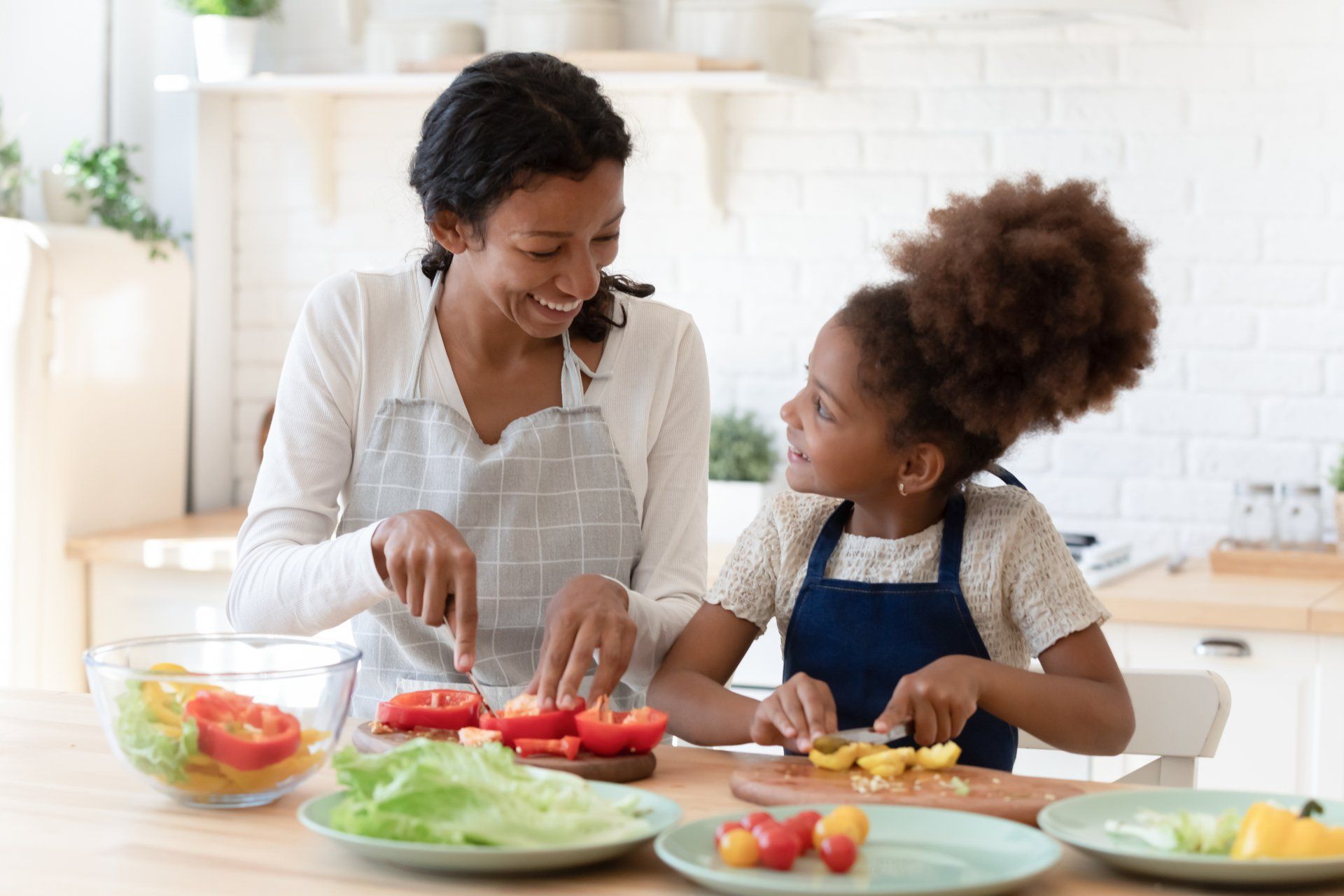 A smiling woman and young girl of color are cooking together in a bright kitchen, cutting vegetables.