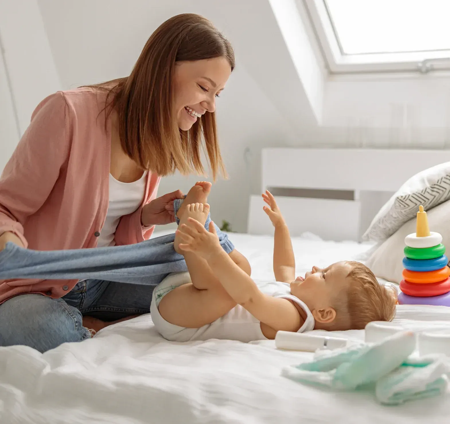 Mother changing baby's diaper on a bed. She's smiling, holding the baby's feet, with toys and supplies nearby in a bright room.