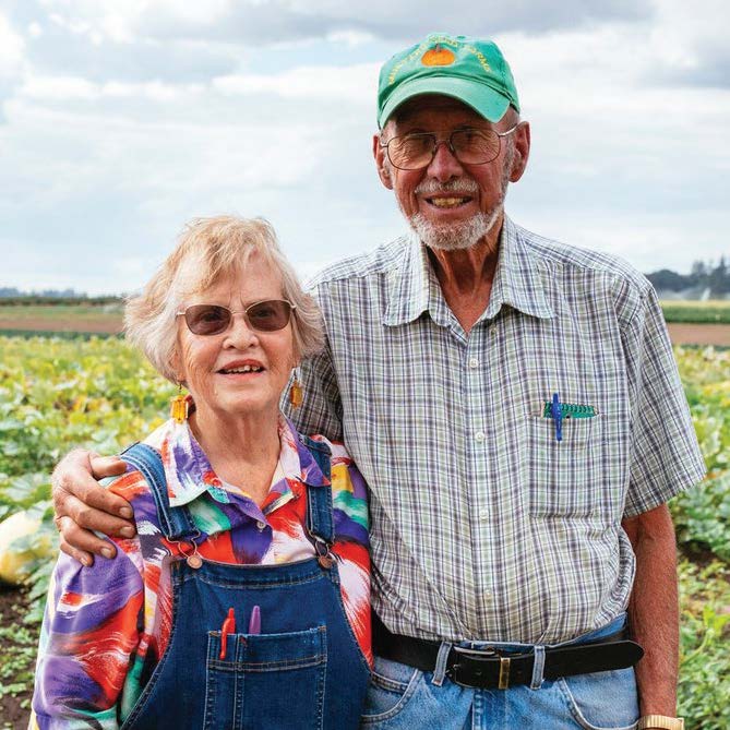 Elderly couple smiles, embracing in a field. Man wears a cap and checkered shirt, woman overalls, both facing the camera.