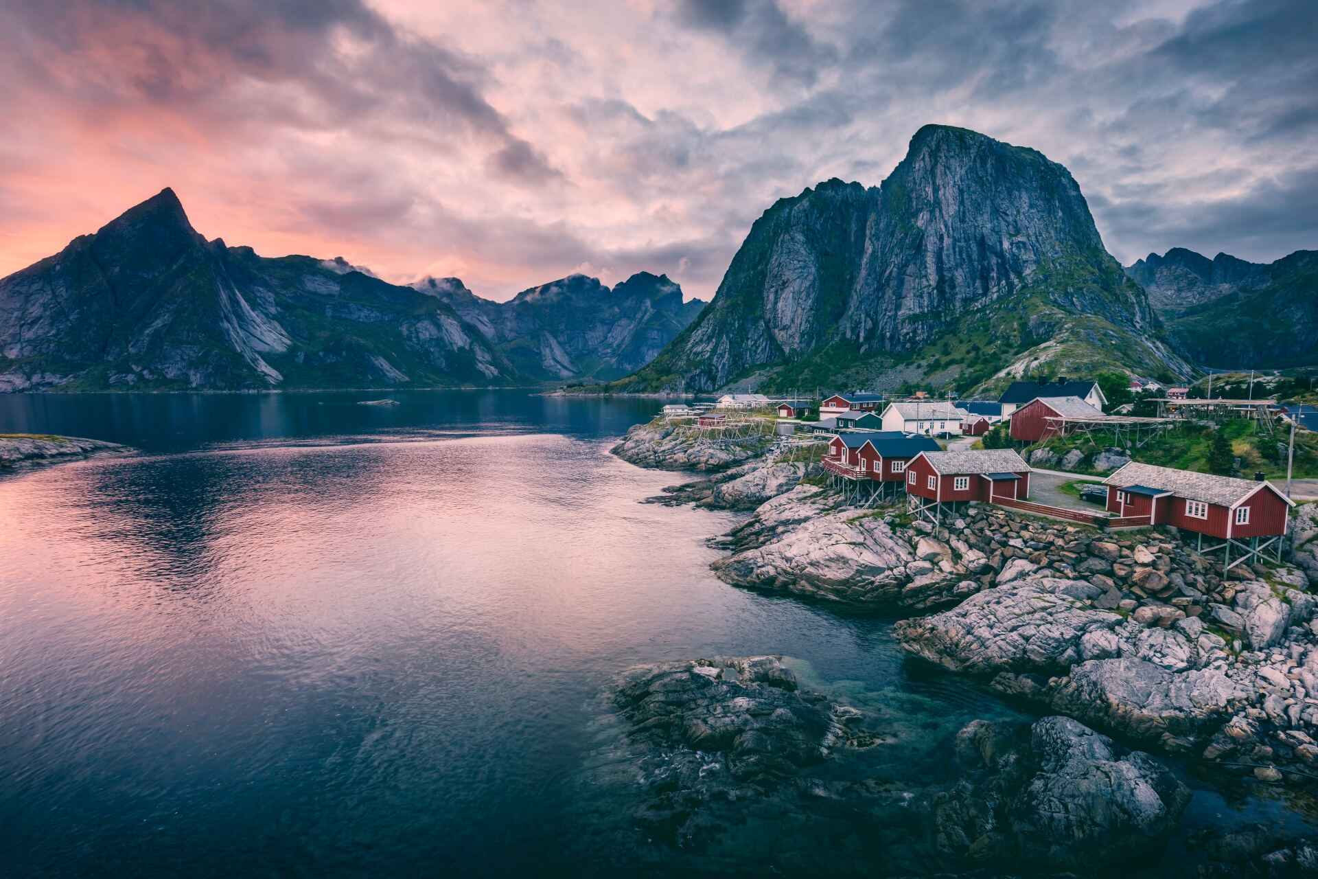 Coastal village with red cabins nestled on rocky shore under mountains at dusk.