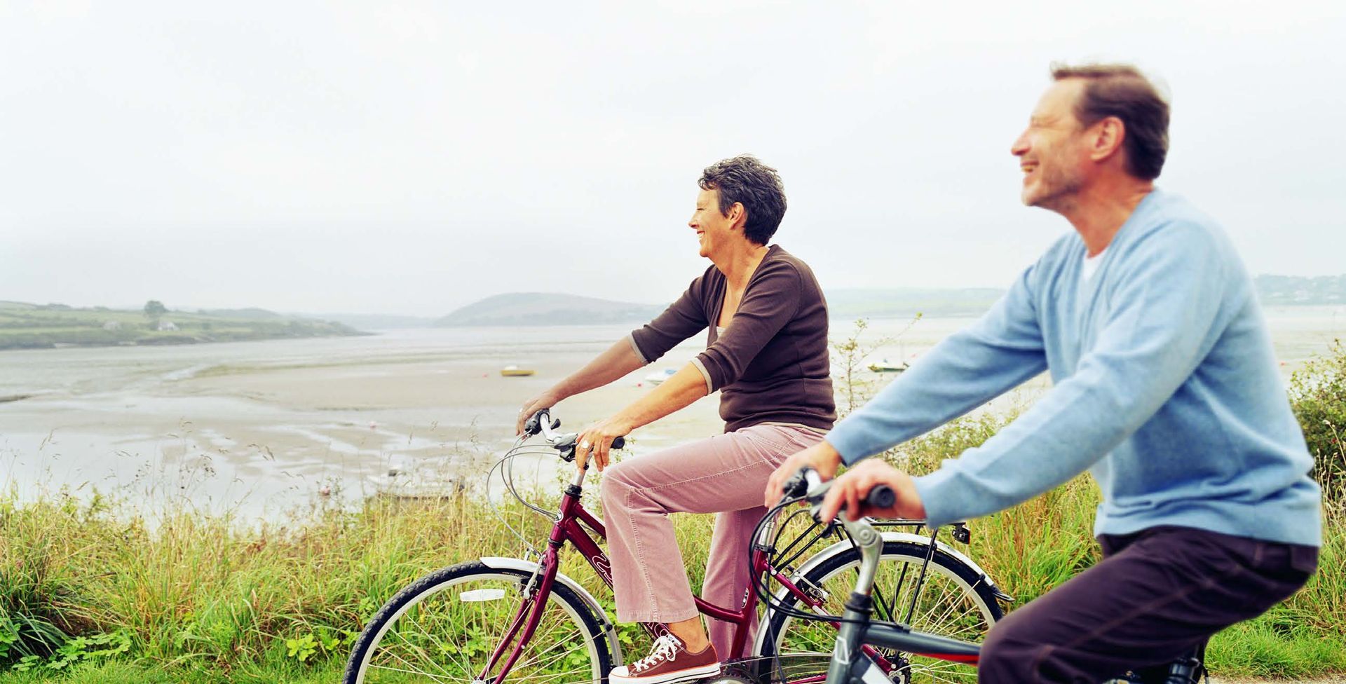 Two people ride bicycles along a path next to a body of water. The woman smiles while looking left; the man smiles, looking up.