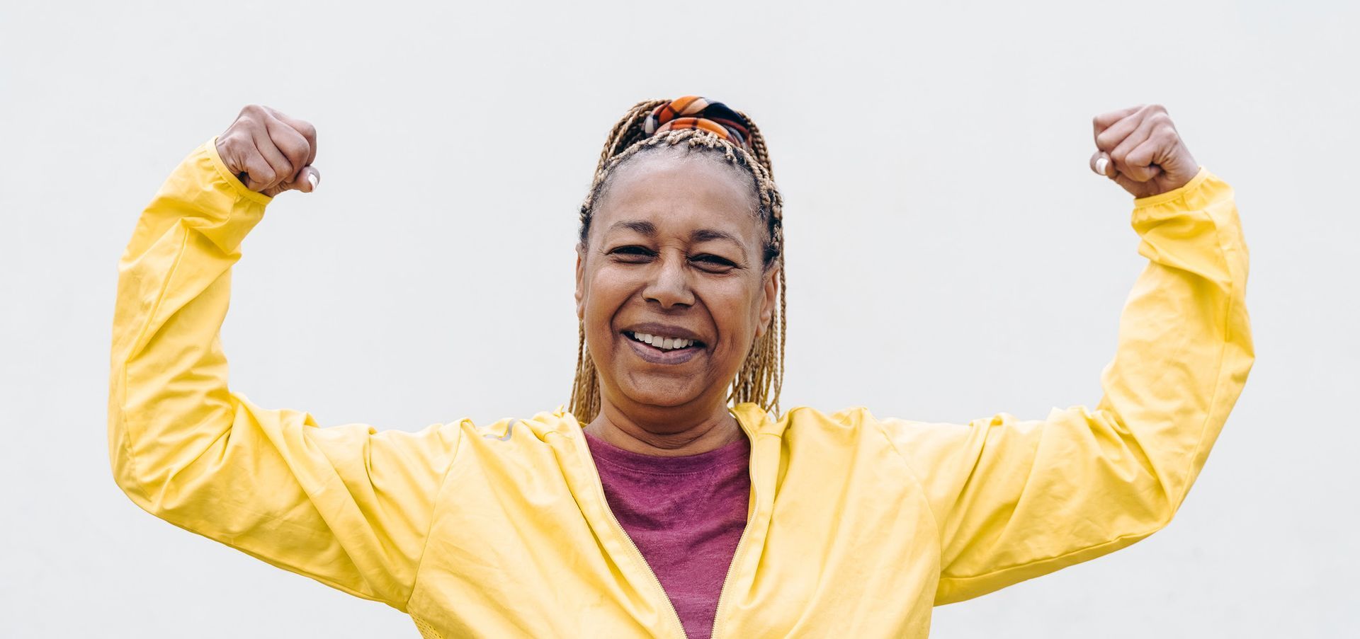 An older woman with blonde braids flexes her biceps with a smile, wearing a yellow jacket and maroon shirt against a white backdrop.