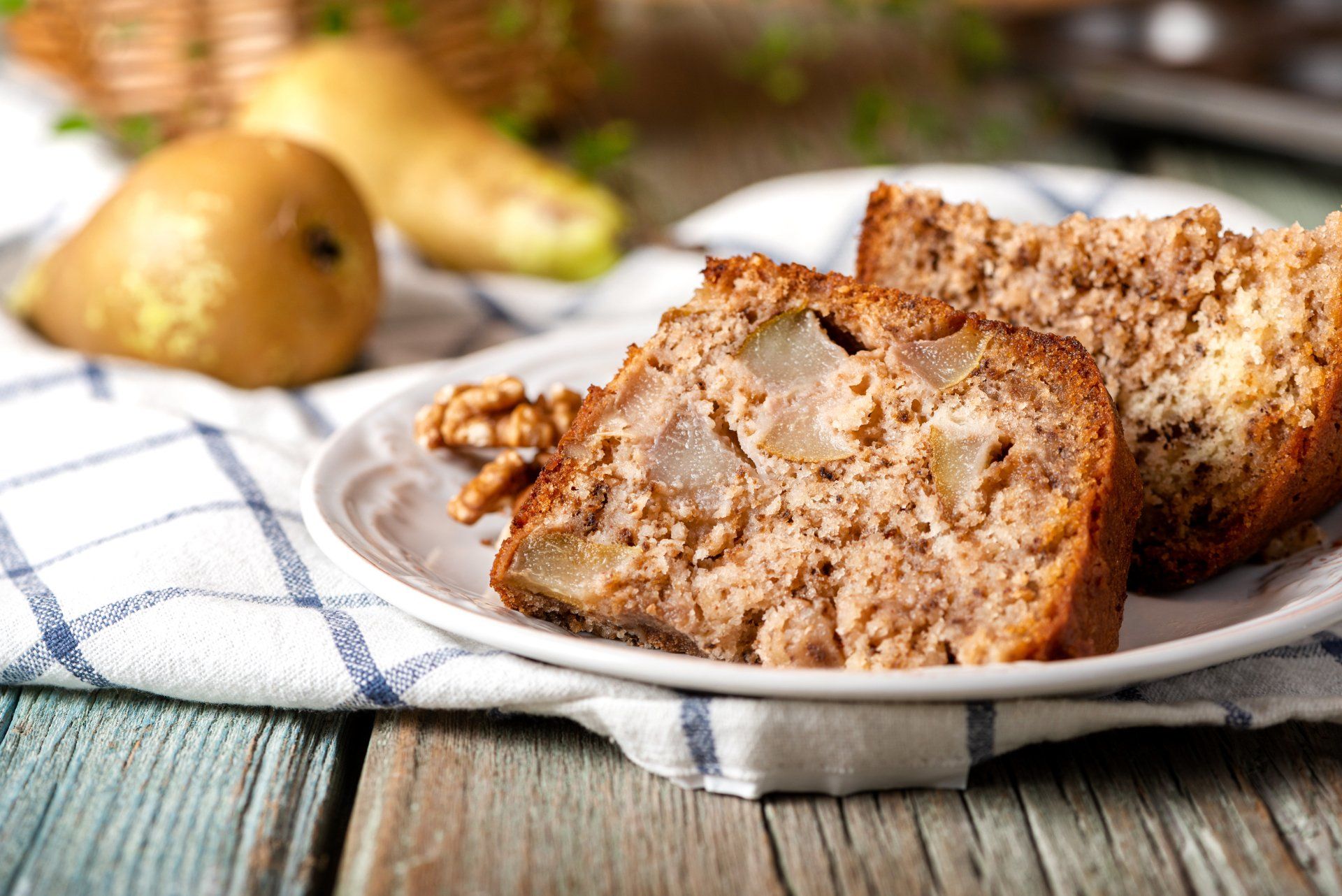 Pear cake slices on a plate with walnuts, pears, and a checkered cloth on a wooden table.