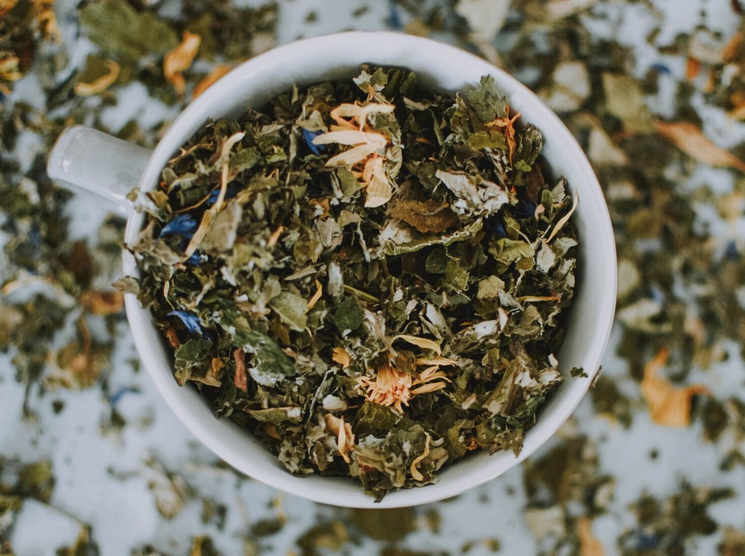 Close-up of a white mug filled with loose-leaf herbal tea with green, orange, and blue flower petals, scattered on a white surface.