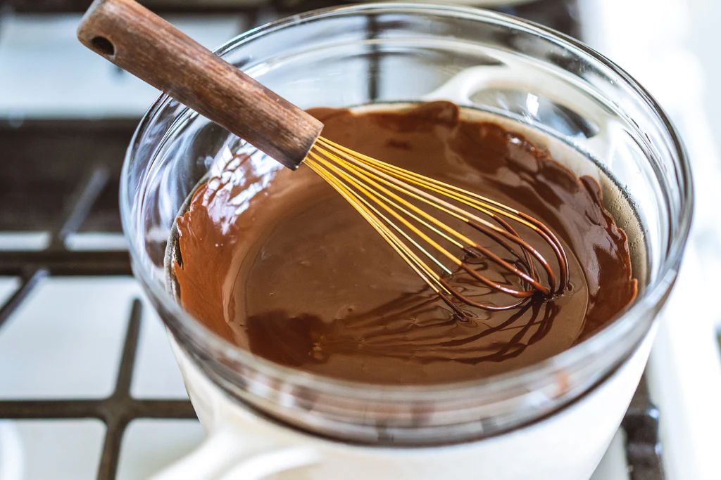 Melted dark chocolate in a glass bowl being whisked with a wooden-handled whisk, resting in a double boiler on a stovetop.