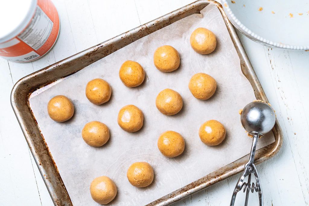 Cookie dough balls on parchment paper-lined baking sheet, ready for the oven. A scoop and container sit nearby.