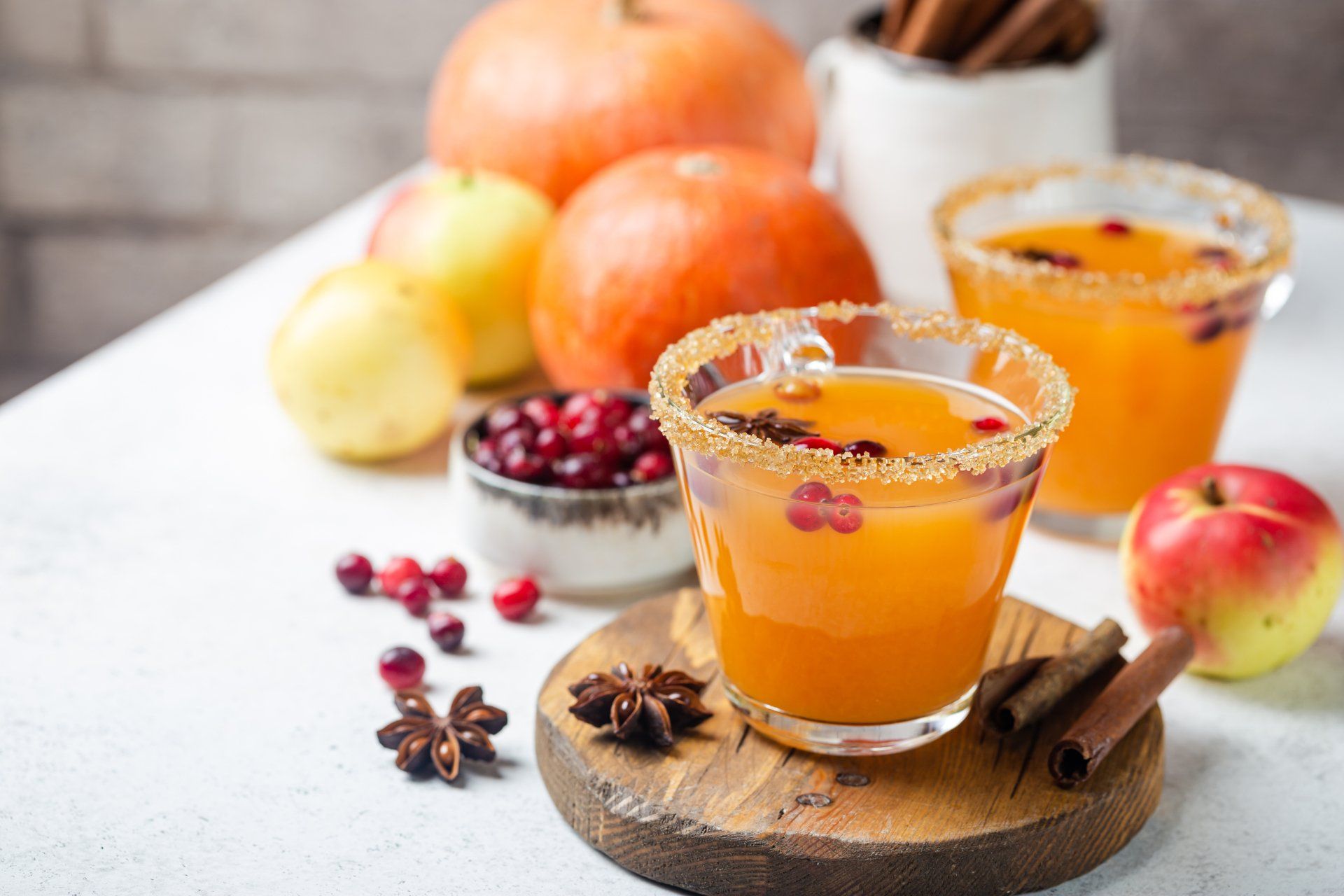 Two pumpkin cider cocktails with sugared rims, surrounded by pumpkins, apples, cranberries, and spices.