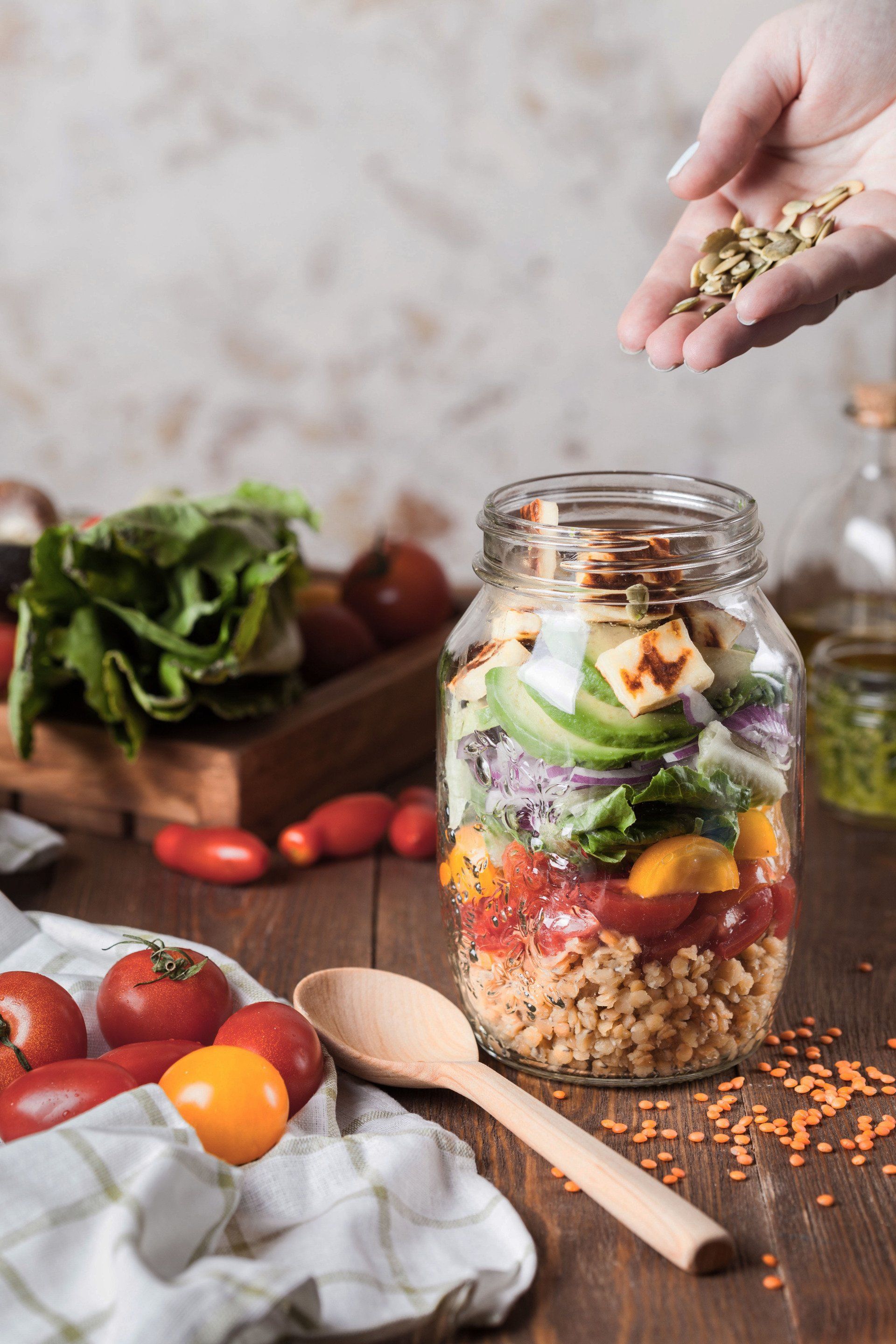 A person sprinkling lentils onto a layered salad in a mason jar on a wooden table, surrounded by ingredients.
