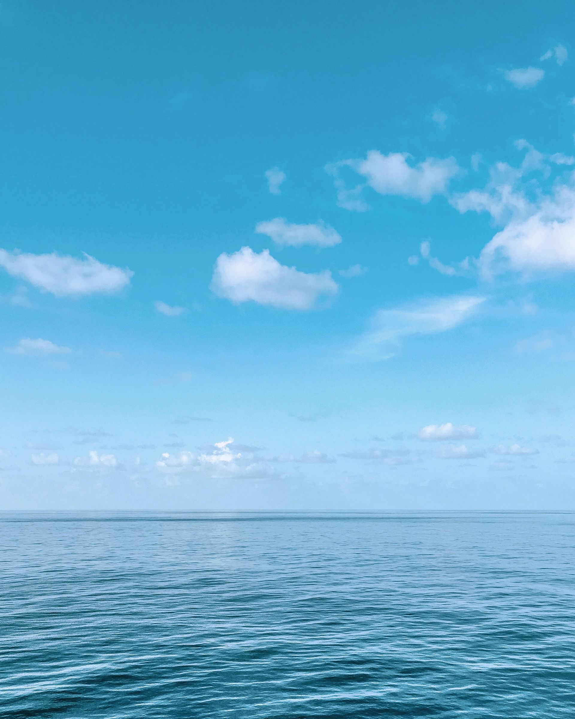 Blue ocean meets a light blue sky with scattered white clouds; peaceful and serene.