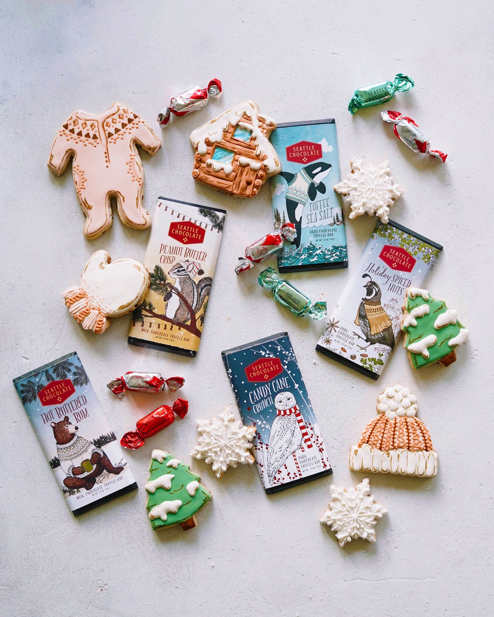 Christmas cookies, wrapped chocolate bars, and candy scattered on a white surface. Includes gingerbread house, sweater, snowflake, and tree cookies, with red and green candies.