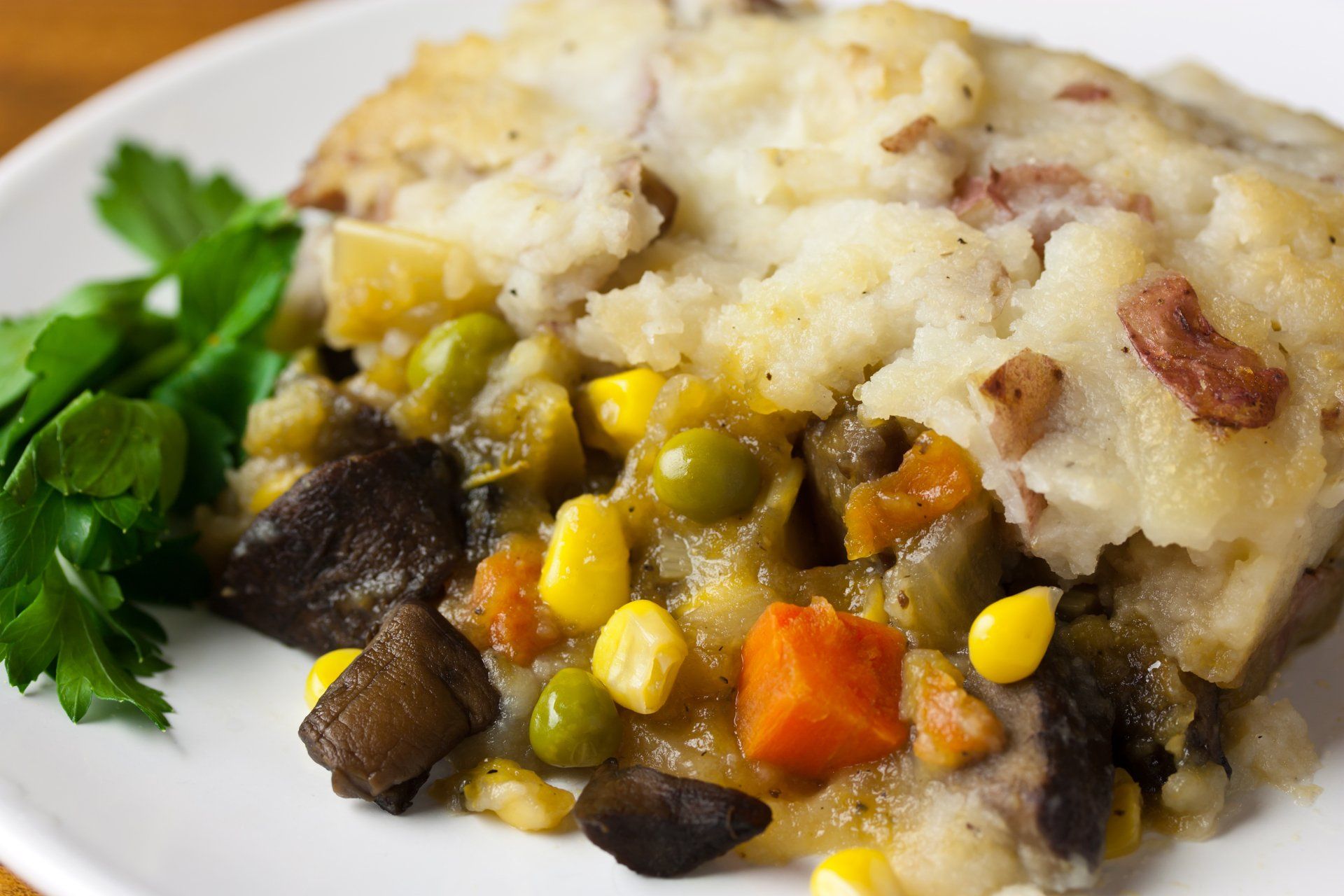 Shepherd's pie on a white plate with visible corn, peas, carrots, and mushrooms, topped with mashed potatoes.