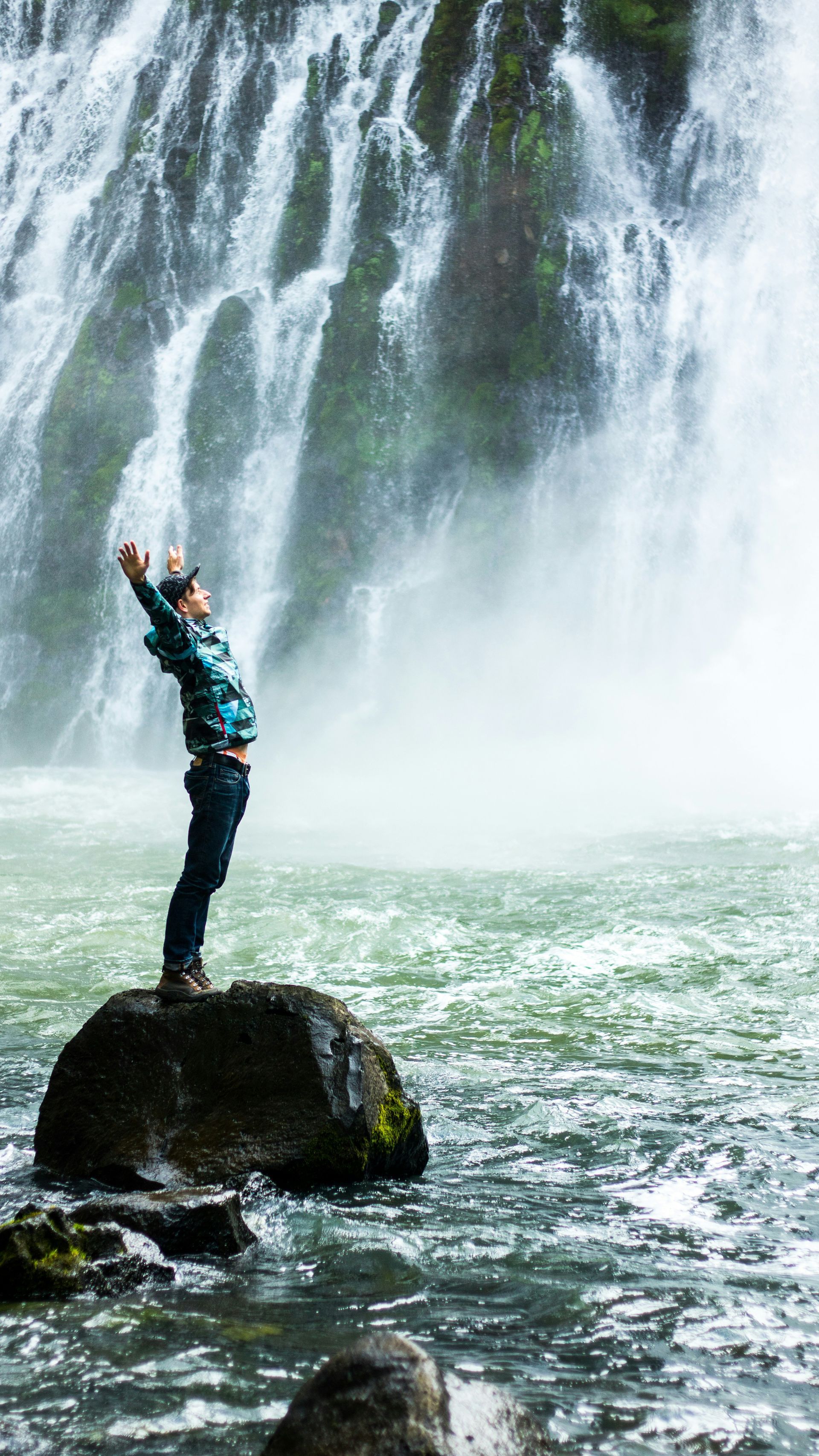 Man with arms raised stands on a rock in front of a waterfall. He wears a blue jacket and a cap in a natural setting.