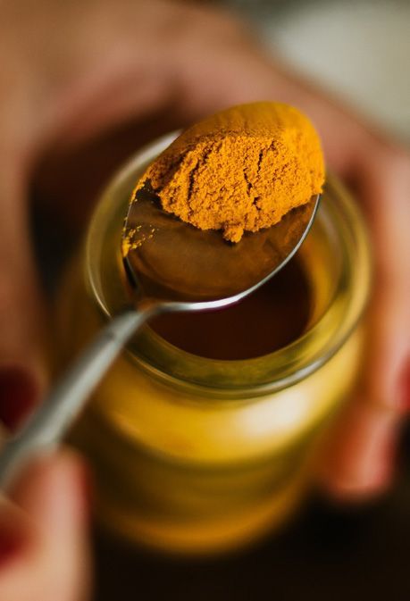 A person's hands holding a spoon filled with bright yellow turmeric powder above a small jar of turmeric.