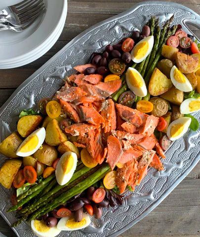 A silver platter with smoked salmon, asparagus, roasted potatoes, hard-boiled eggs, olives, and cherry tomatoes arranged decoratively.