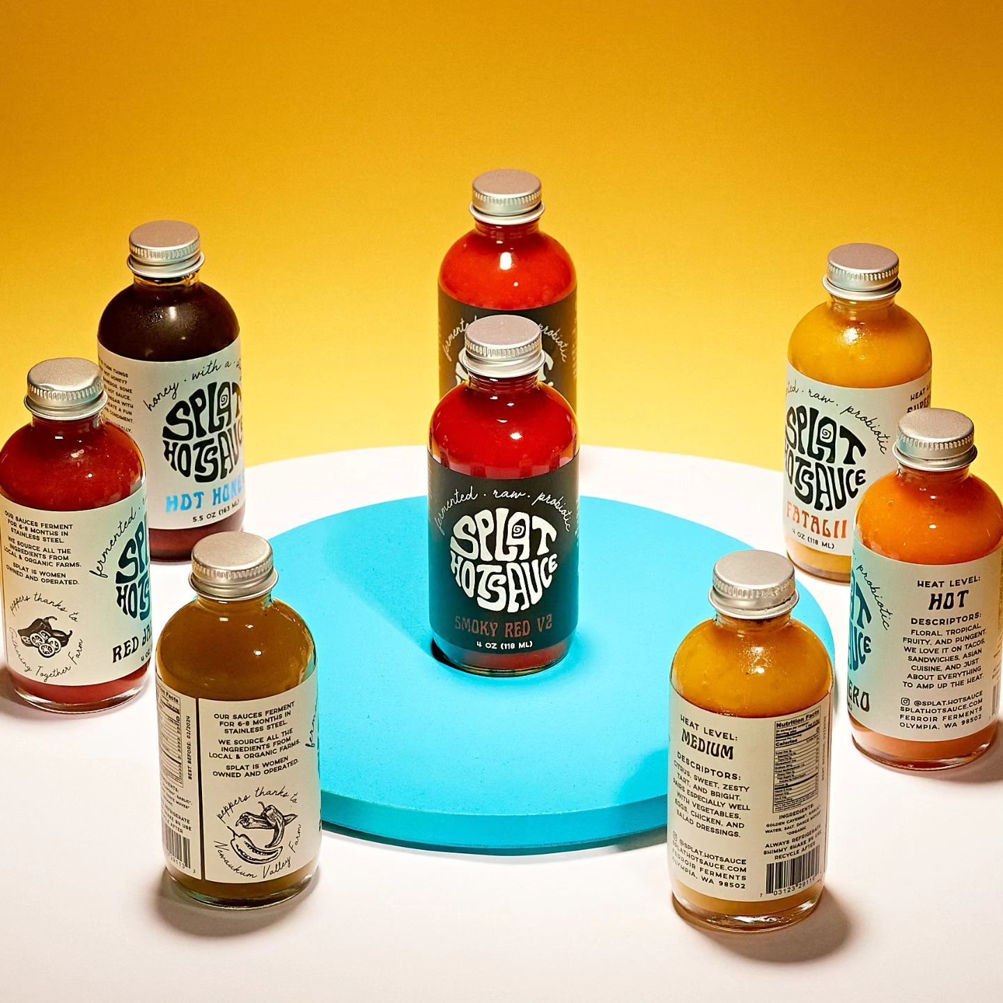 Eight bottles of colorful hot sauce arranged on a blue and white surface against a yellow background.
