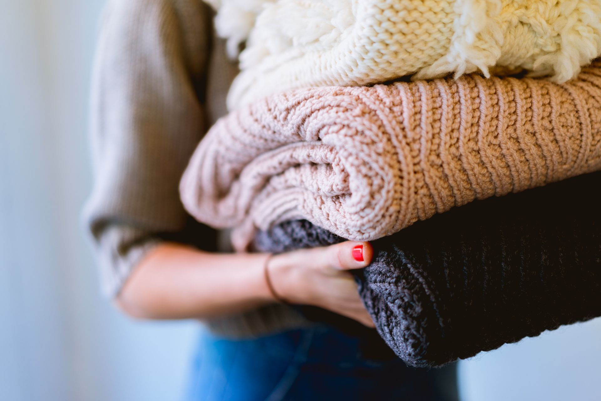 Woman holding a stack of folded sweaters in various neutral colors, including white, pink, and black.