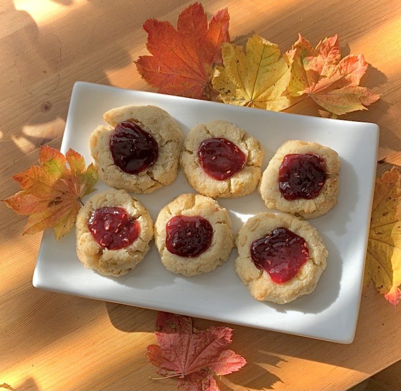 Six thumbprint cookies with jam centers on a white plate, autumn leaves around.