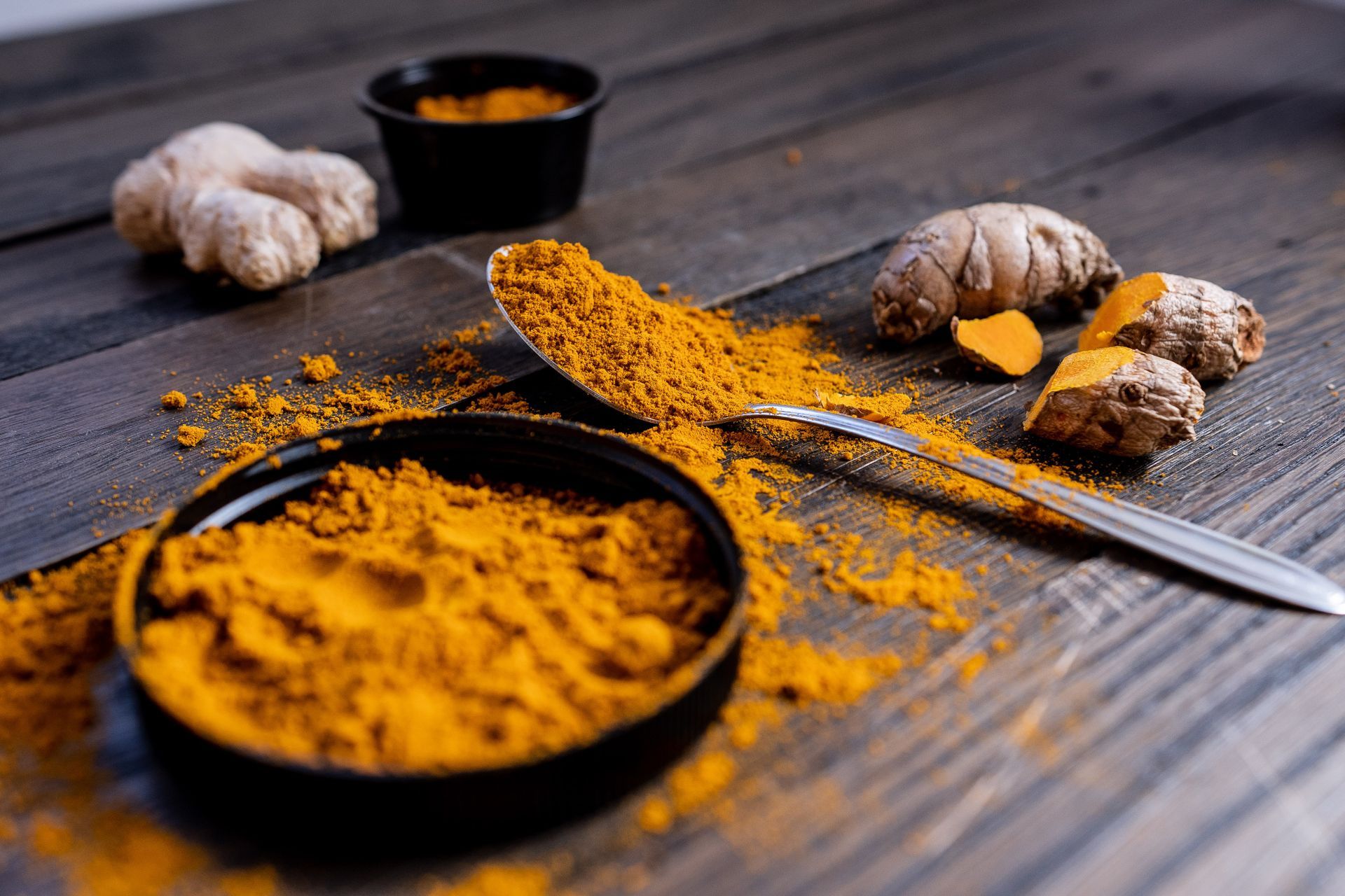 Turmeric and ginger root with turmeric powder in a small black dish and on a spoon, scattered on a wooden surface.