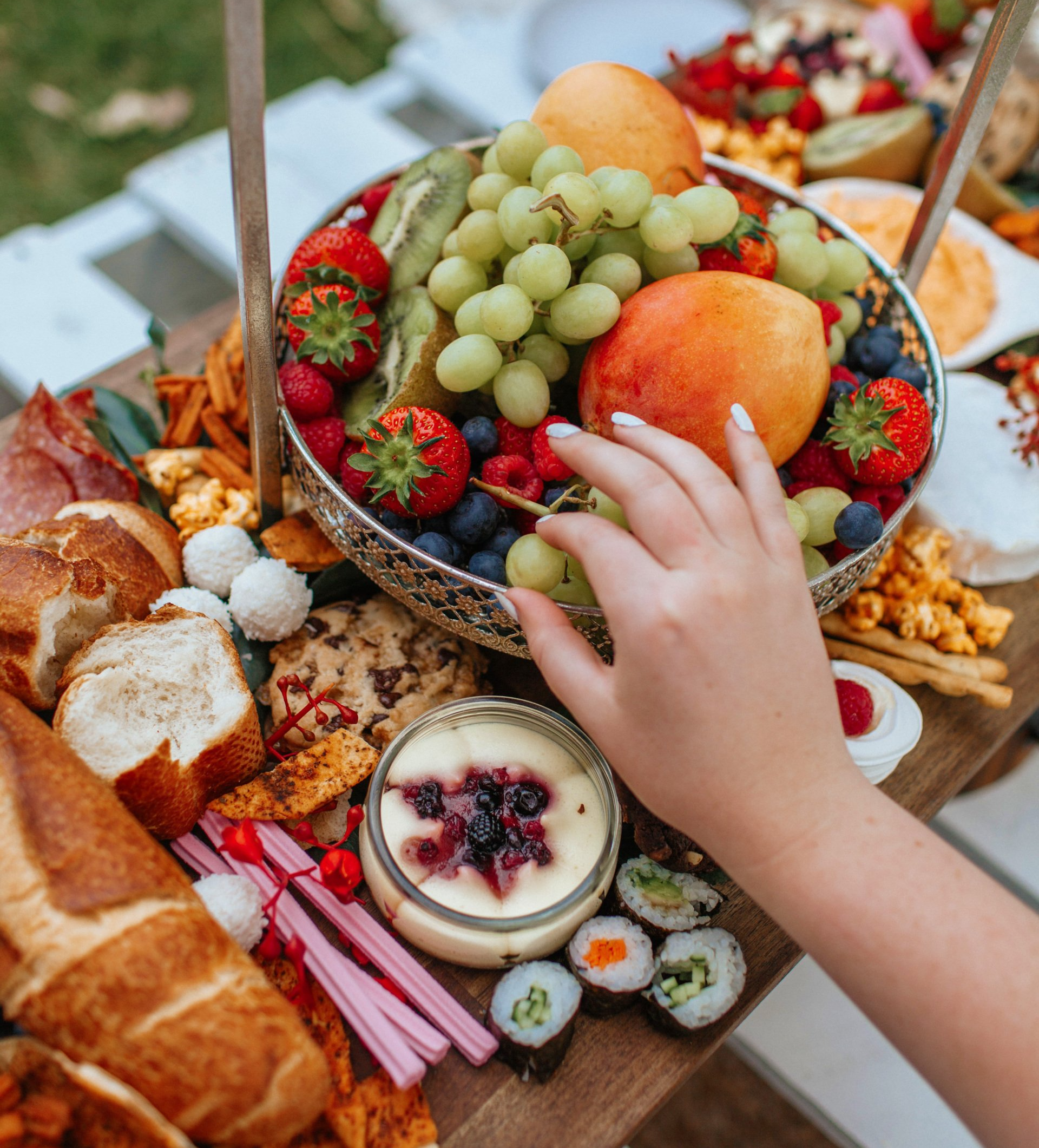Close-up of a charcuterie board with fruit, cheese, and other snacks. A hand reaches for grapes from a silver bowl of fruit.