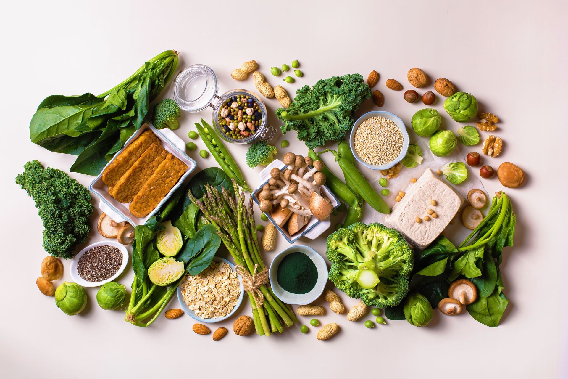 Overhead view of various plant-based foods, including greens, vegetables, nuts, and seeds arranged on a light-colored surface.