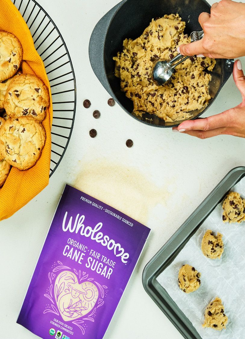 Woman scooping cookie dough from a bowl. Chocolate chip cookies on a baking sheet and in a basket; Wholesome cane sugar bag visible.
