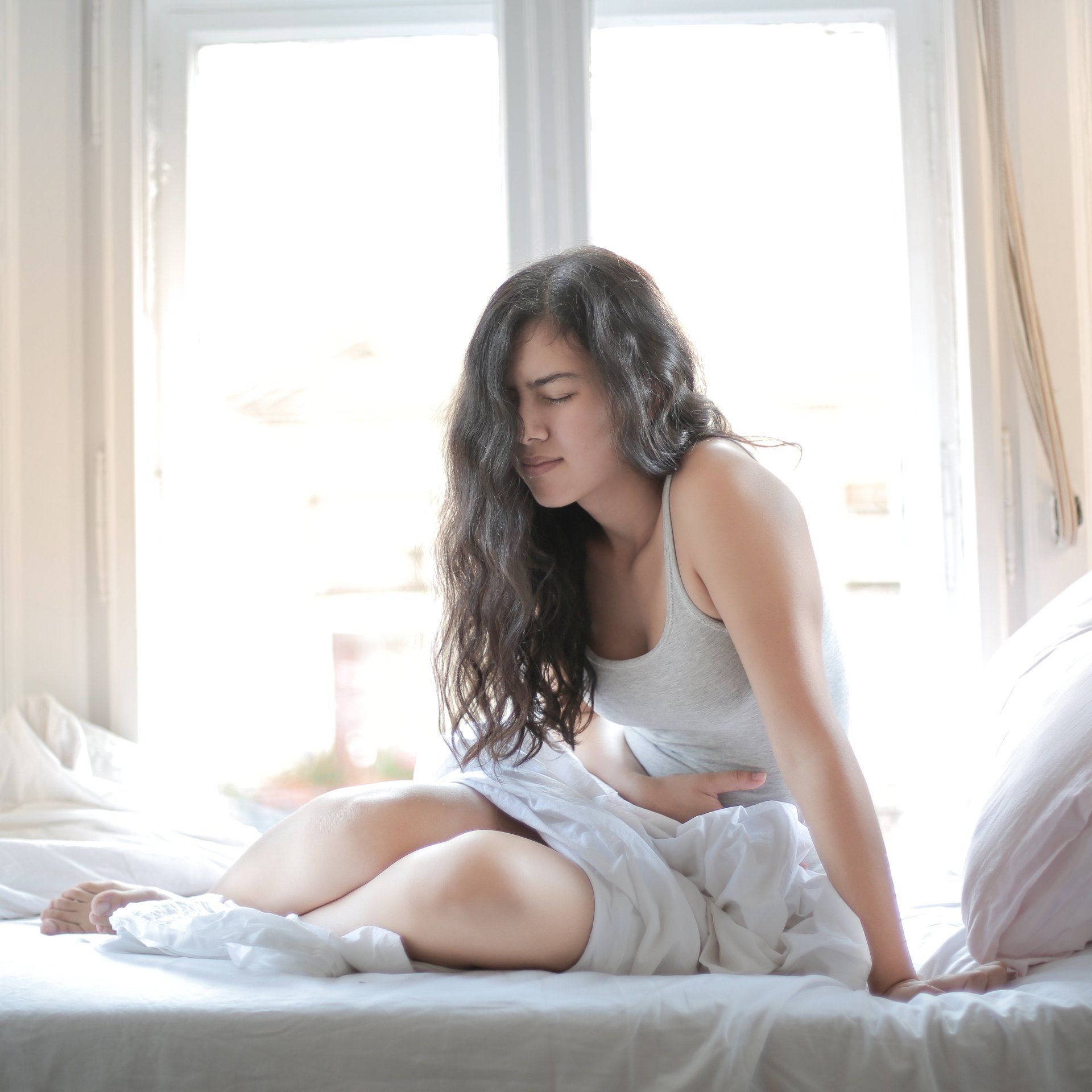 Woman sitting on a bed, clutching her abdomen in pain. She has long dark hair and is in a bright room, with a window behind her.