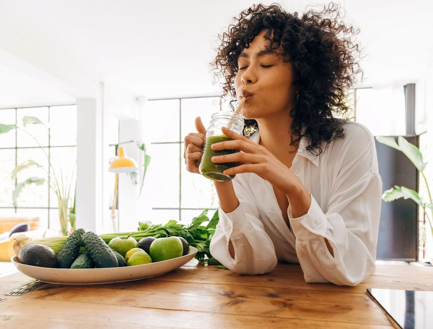 Woman in white shirt sips a green smoothie with a straw, surrounded by fresh produce on a wooden table in a bright kitchen.