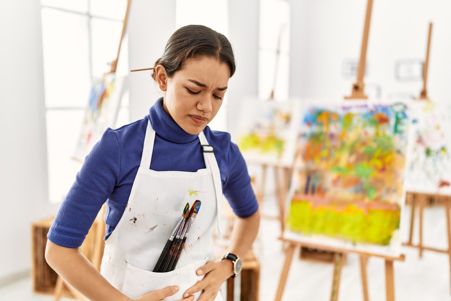 Woman in an art studio clutching her stomach in pain, wearing an apron with paintbrushes. Paintings on easels are in the background.