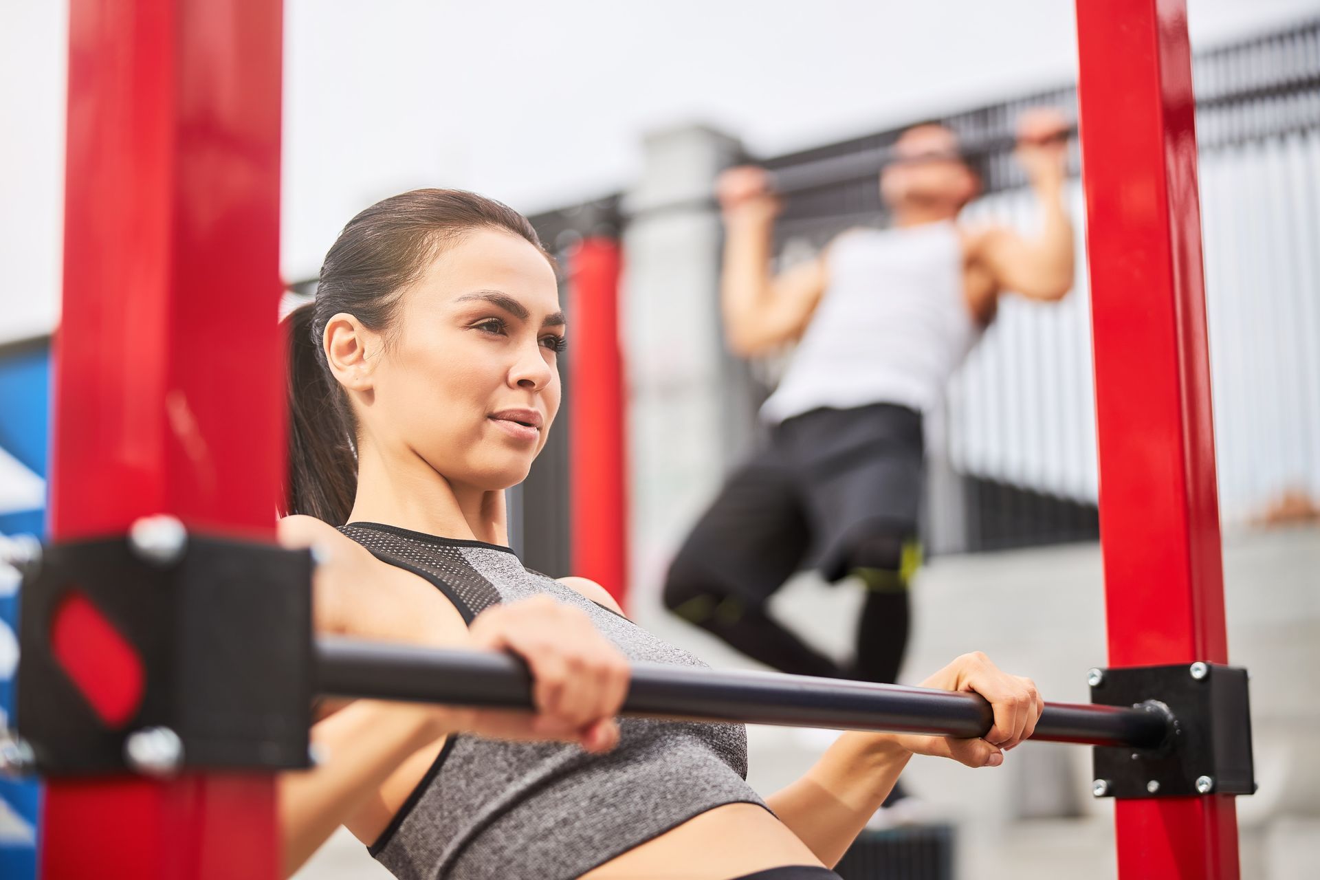 Woman doing a pull-up, looking focused, at an outdoor fitness park; another person in the background is also exercising.