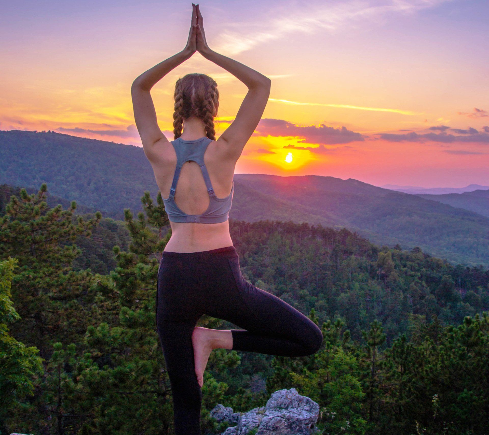 Woman in yoga pose on a mountain, arms raised, facing a colorful sunset.